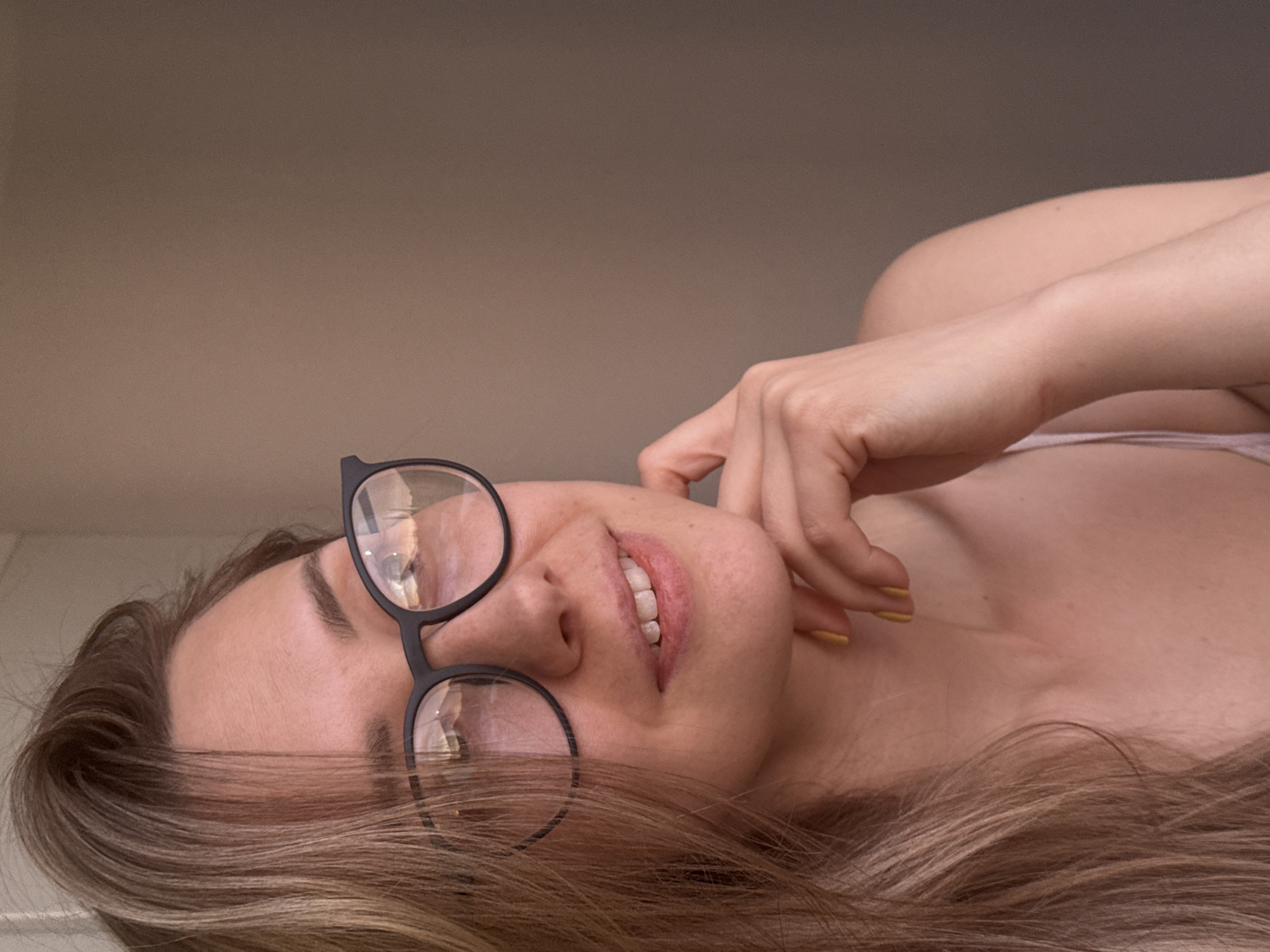 A fair-skinned woman with light brown hair is lying on her back, with her head tilted slightly to her left. She is wearing black-framed glasses, and her right hand is near her face. She is smiling and shows teeth. She is located indoors. There is no information available regarding the location of the scene in the city of Chaikovsky, Russia.