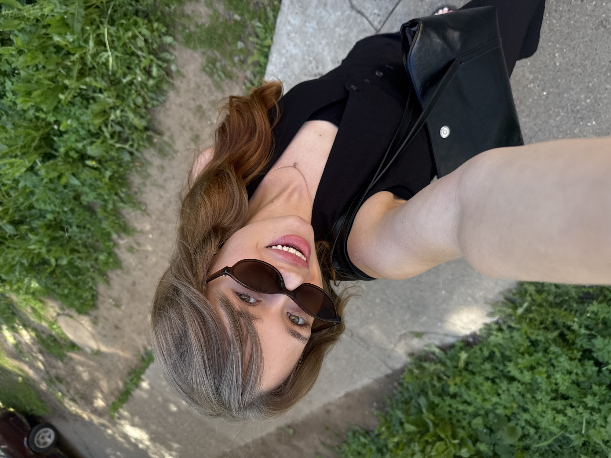 The image is a high-angle, selfie-style shot of a young woman standing outdoors, likely in a suburban or semi-rural area of Chaikovsky, Russia. She has medium-length, wavy auburn hair with lighter streaks and is wearing dark sunglasses, a black sleeveless top over a black tank top, and a delicate gold necklace. She is smiling and holding a black shoulder bag.
The environment consists of a paved pathway or sidewalk on the right, bordered by lush green grass and foliage on the left. Sunlight casts dappled shadows on the ground, suggesting a sunny day. In the background, a portion of a dark-colored car and more greenery are visible. The overall impression is that of a casual outdoor moment captured on a bright day.