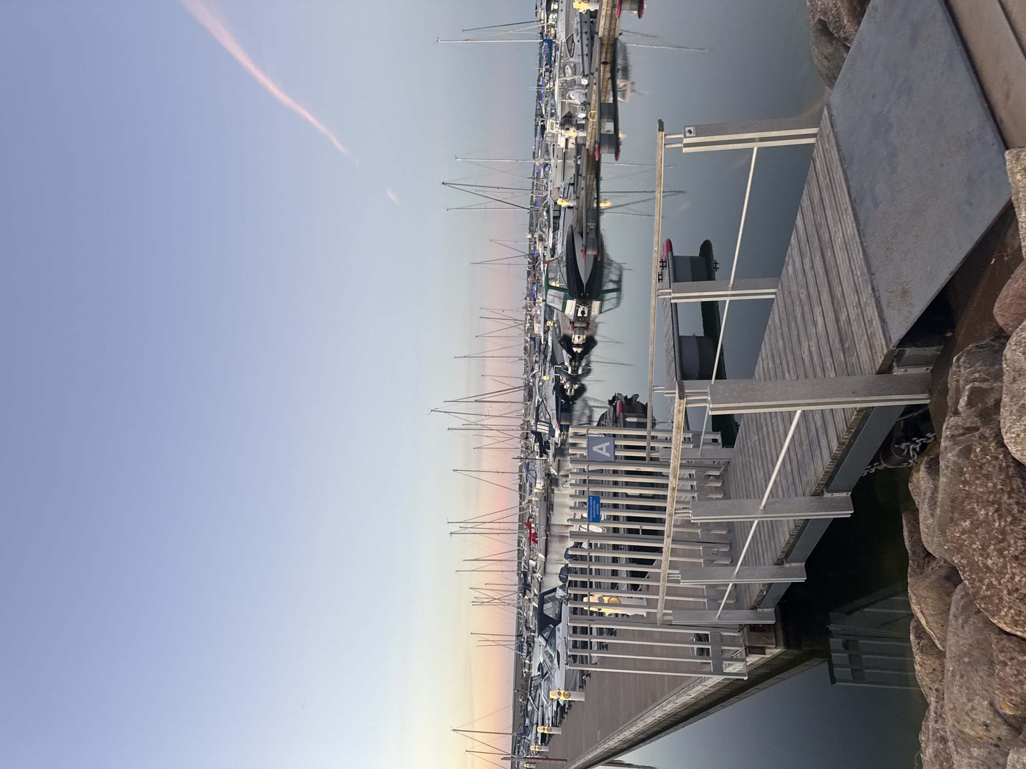 The image shows a marina in Tallinn, Estonia, with numerous sailboats and motorboats docked. The foreground features a wooden pier with metal railings and a ramp leading down to the water. The sky is a soft gradient of pale blue and pink, with a faint pink cloud streak. The water is calm and reflects the boats and the sky. The scene appears to be set during either sunrise or sunset. There are no people visible in the image.