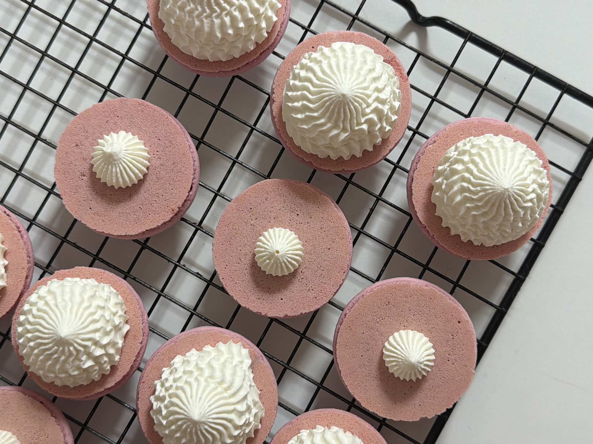 The image shows a top-down view of several pink macarons arranged on a black wire cooling rack. The macarons are of varying degrees of decoration; some are topped with a large swirl of white cream filling, while others have only a small dollop of cream in the center. The rack is placed on a white surface. The image does not contain sufficient information to place the location of this scene in Tel-Aviv, Israel. There are no identifiable people, actions, or interactions occurring.