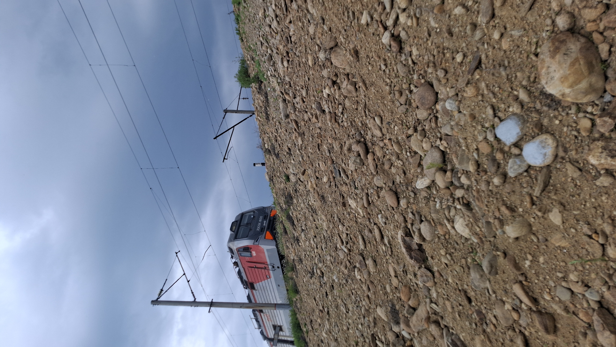 An overhead shot captures a Russian train, specifically a 33CSK 1600 model, traveling on a rocky embankment under a cloudy sky. The train is grey and red, with the number 1600 visible on its side. Overhead power lines stretch across the frame, supported by tall poles. The embankment is covered in a dense layer of brown and grey stones of various sizes, with sparse green vegetation peeking through. The sky is a mix of dark grey and lighter blue clouds, suggesting an overcast day. There are no people visible in the image.