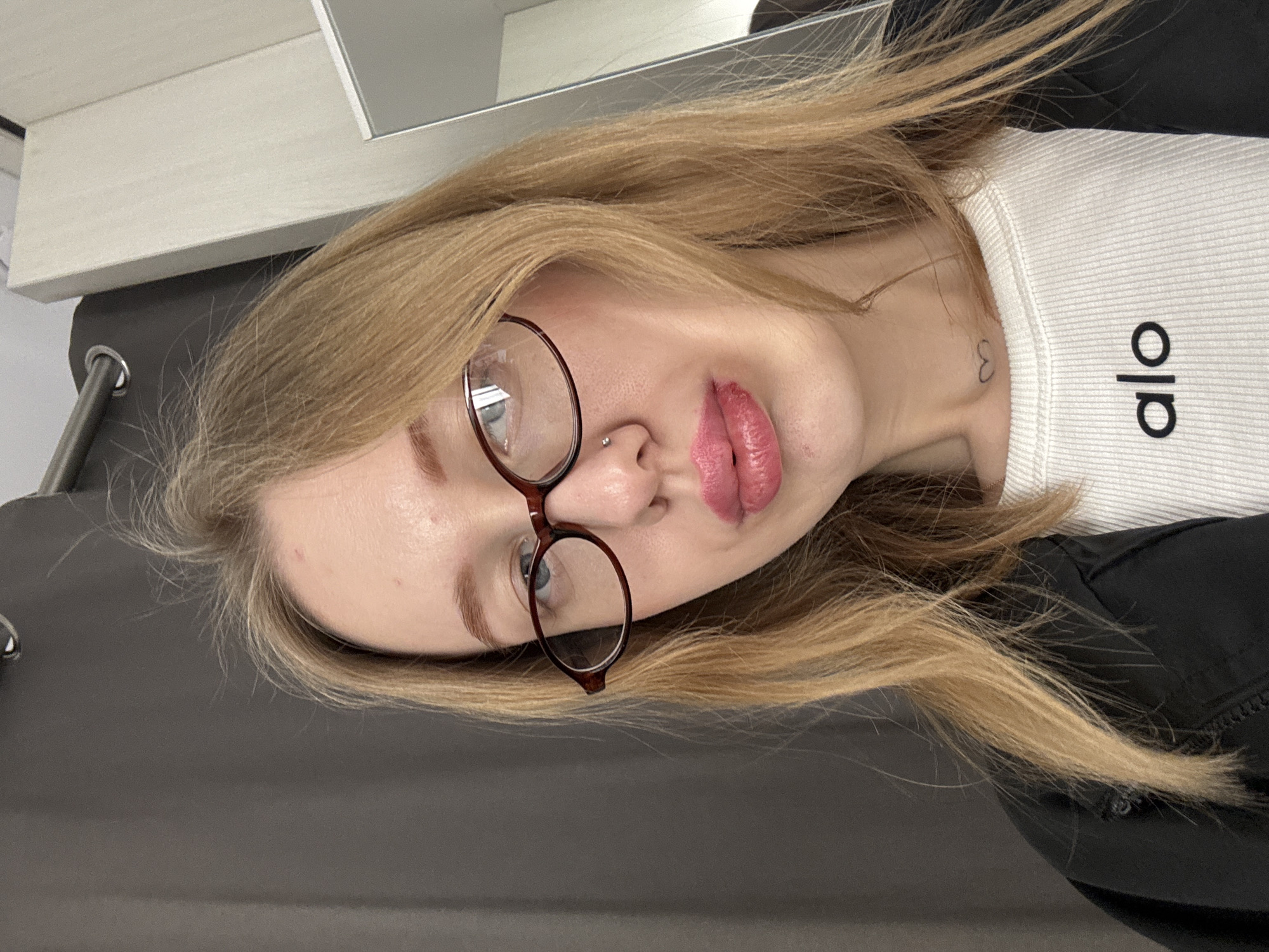 A direct, front-facing selfie captures a young adult female from the chest up, positioned indoors in Rostov-on-Don, Russia. She has fair skin, long, straight light brown hair parted centrally, and blue eyes partially obscured by dark brown, oval-framed glasses. A small silver stud pierces her right nostril. Her lips are full, coated in pink lipstick, with a visible mark or irritation on the lower lip. Small red blemishes are present on her forehead. A small, black outline of a heart is tattooed on the front of her neck. She wears a white ribbed top displaying "alo" in black text, layered beneath a dark-colored jacket or shirt. The background features a dark grey curtain on the left, suspended from a metal rod with a visible silver eyelet. To the right, a light-colored, possibly white or beige, wall or cabinet unit is partially visible, with a reflective surface (likely a mirror) on the far right edge. The lighting is consistent with an indoor setting.