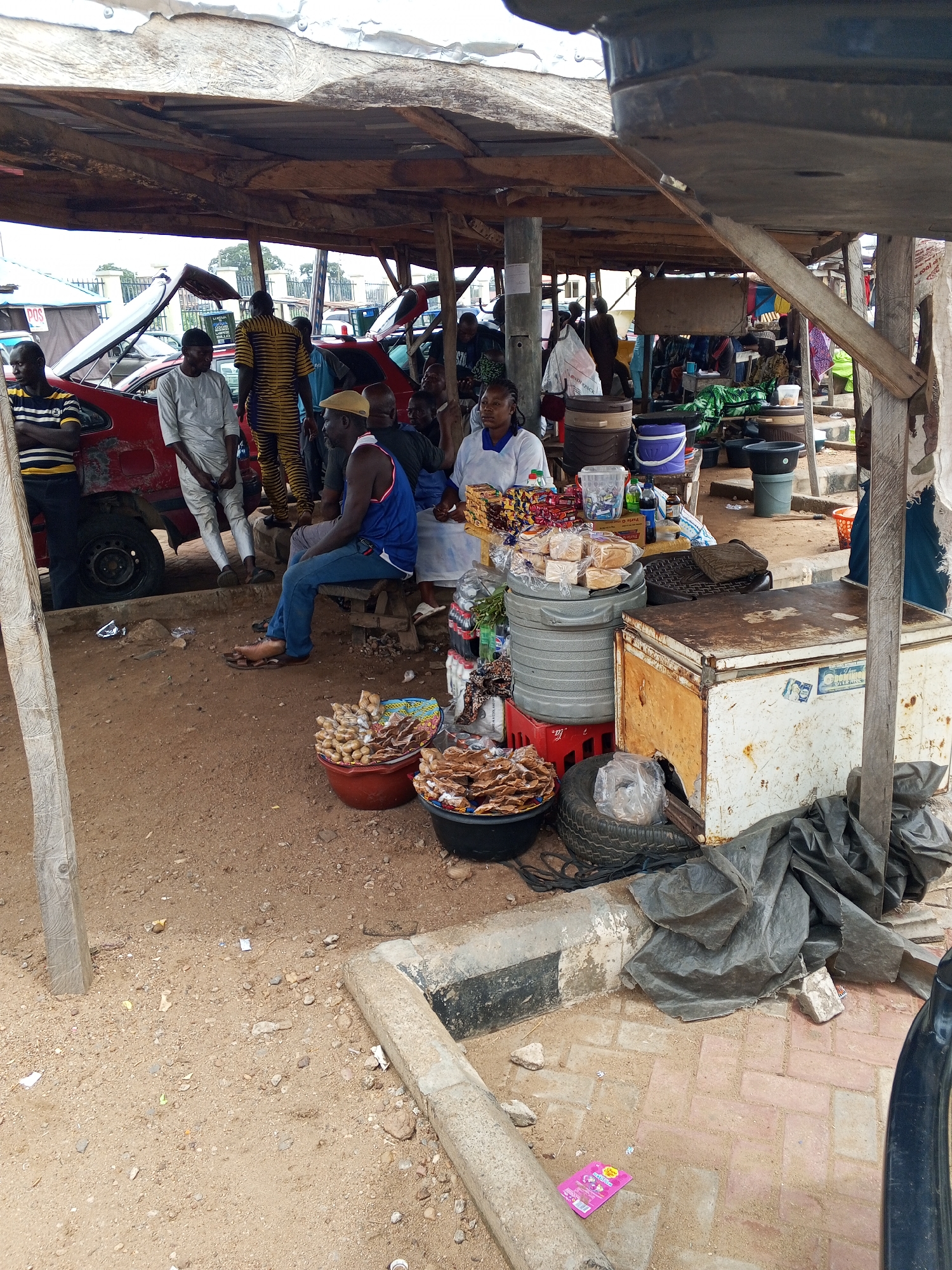A bustling scene unfolds at an outdoor market stall in Lokoja, Nigeria. A canopy roof supported by wooden posts shades the area, offering respite from the elements. Several individuals are present, engaged in various activities. Some are seated, while others stand around a red car with its hood open, possibly discussing repairs or maintenance.
The market stall itself is laden with goods, including stacked bread, packaged snacks, and drinks. Buckets overflow with what appears to be a type of dried food. A worn-out refrigerator and plastic containers sit adjacent to the food items.
The ground is primarily dirt, with a paved section near the stall's edge. Litter is scattered about, adding to the atmosphere of a busy, active marketplace.