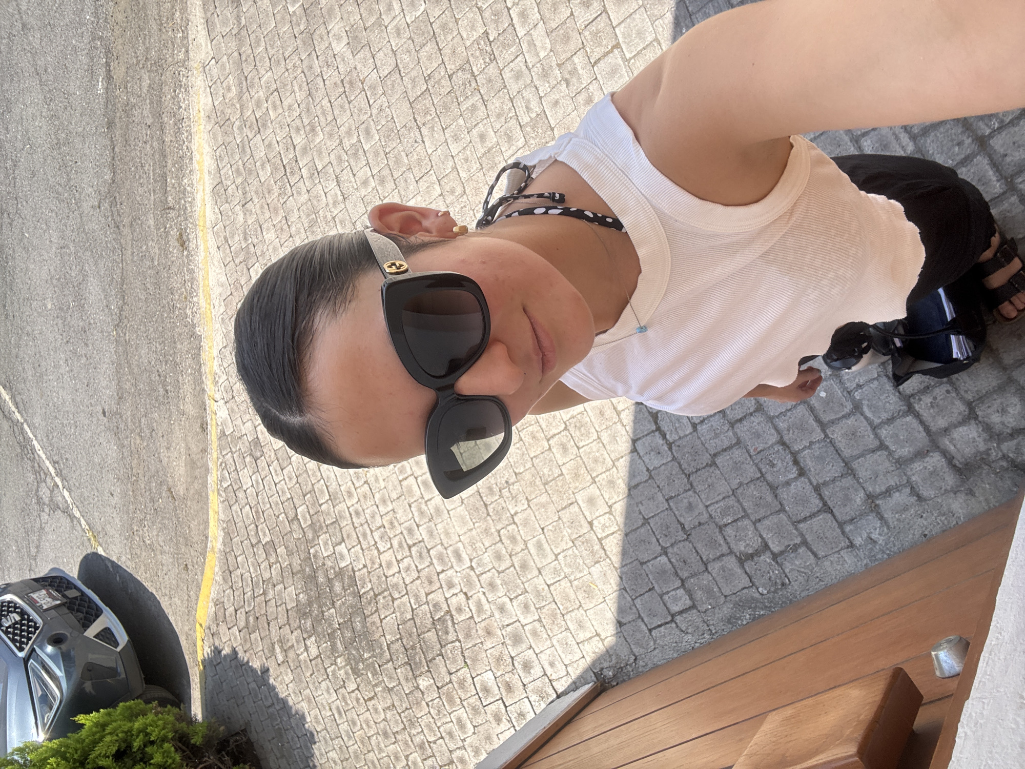 The image captures a high-angle, top-down view of an individual in San Bernardino Tlaxcalancingo, Mexico, under bright sunlight. The main subject, a person with dark hair pulled back, is looking up towards the camera. Their face is partially obscured by large black cat-eye sunglasses, which feature gold-colored interlocking 'G' logos (likely Gucci) on the temples. A small gold-colored hoop earring is visible in their right ear.
The person is wearing a white ribbed tank top, black pants or shorts, and black sandals with thick straps. A black and white polka-dotted strap, possibly part of swimwear or another top, is visible near their shoulder, along with a delicate necklace holding a small blue bead.
The setting is an outdoor paved area. The ground beneath the person is covered in light-colored cobblestones, transitioning to a darker asphalt or concrete road with a yellow painted line on the left side of the frame. Strong shadows are cast across the ground, indicating a sunny day, likely mid-day or early afternoon. In the background on the left, the front section of a dark grey car, possibly an SUV, is partially visible, parked next to some green shrubbery. The bottom right corner of the image reveals a wooden surface, suggesting a deck, bench, or outdoor furniture, with a small metallic fixture resting on it. The individual appears to be casually posing for the photograph.