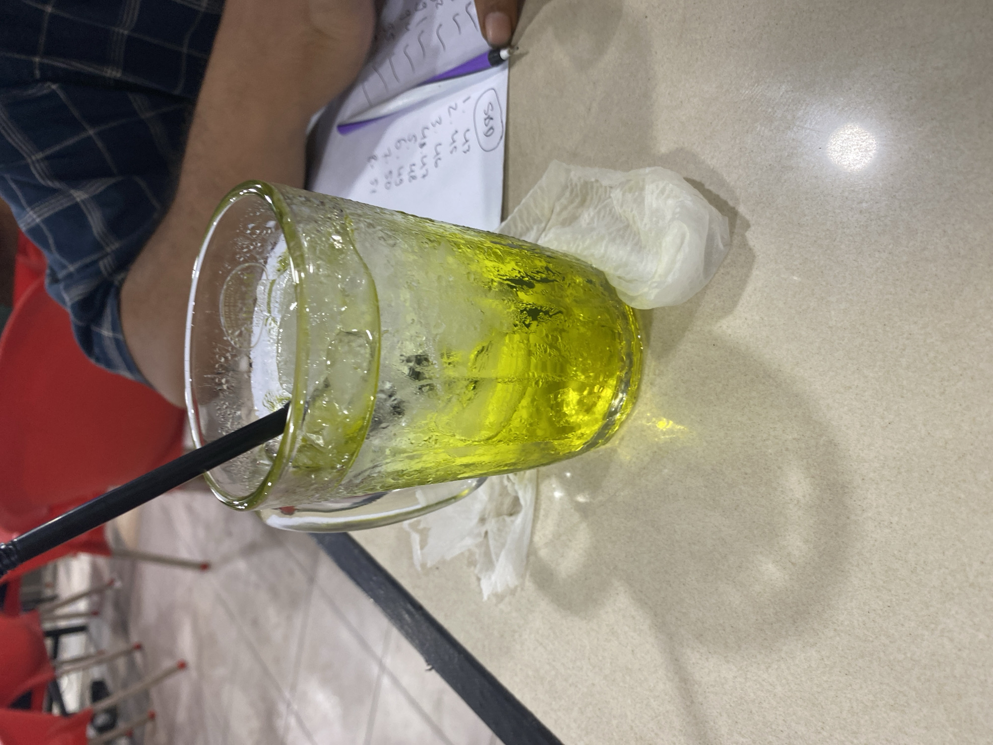 The image shows a close-up, top-down view of a glass of bright yellow liquid with ice, placed on a speckled, light-colored table. A black straw is submerged in the drink. To the left of the glass, a person's arm, clad in a blue and white checkered shirt, is visible, along with a portion of their body. In the background, a notebook with handwritten numbers and symbols is open, and a pen is being held over it, suggesting someone is writing or calculating. A crumpled white tissue is on the table near the glass. The lighting suggests an indoor setting, possibly during the daytime, with a bright light source creating a circular reflection on the table. The overall scene appears casual and ordinary, with no distinct emotional cues. The visible text on the notebook includes numbers such as "43" and a circled "S60". The scene is set in Lhokseumawe, Indonesia.