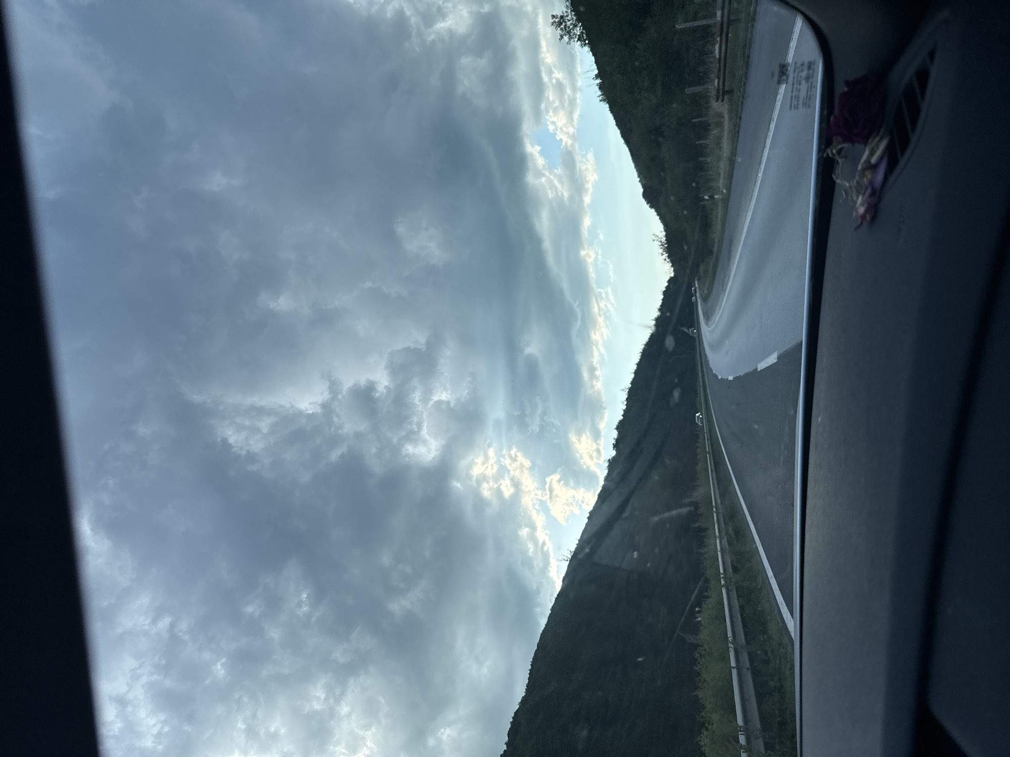A high-angle, wide shot captures a road winding through a mountainous landscape. The sky is filled with dramatic, dark clouds, but shafts of sunlight break through in places. The road, with a white line marking its edge, curves to the right and disappears into the distance. On either side of the road, steep, tree-covered hills rise up, their slopes darkened by the overcast sky. A metal guardrail runs along the side of the road. A dark dashboard with a small cluster of dried flowers and keys is visible in the foreground. The scene appears to be a rural highway in the vicinity of Dolno Varshilo, Bulgaria.