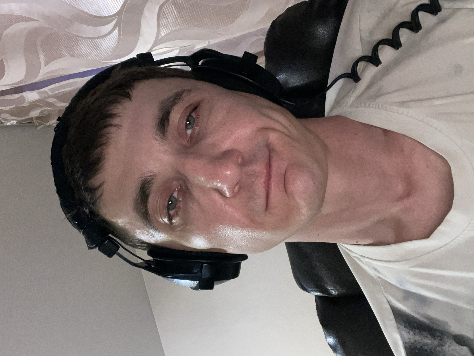 A man with short, dark hair is wearing large, black headphones and a white t-shirt. He is looking directly at the camera with a slight smile. His face appears to be sweaty. He is seated in a black chair in front of a window with sheer curtains. The background is dimly lit. The location is Laishevo, Russia.
