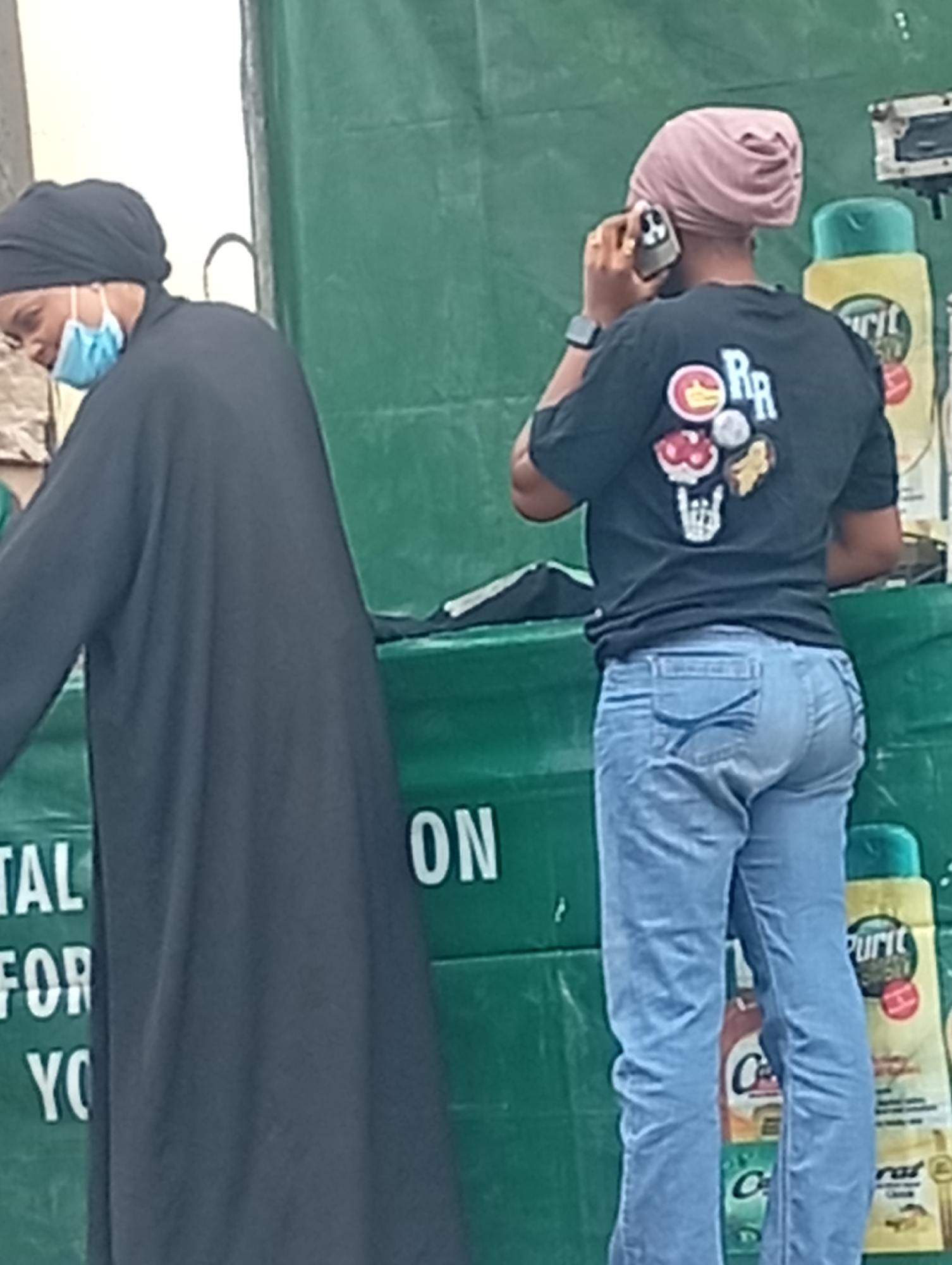 The image depicts two individuals in an outdoor setting. One individual, wearing a black headscarf and face mask, stands near a table with green coverings. The other individual, wearing a pink head covering, a black t-shirt with an emblem, and blue jeans, is holding a phone to their ear. Behind the table are stacked household cleaning products, suggesting a market or commercial venue. A banner with the words "TAL FOR YOU ON" is partially visible. The location of the scene is the city of Maiburiji, Nigeria.