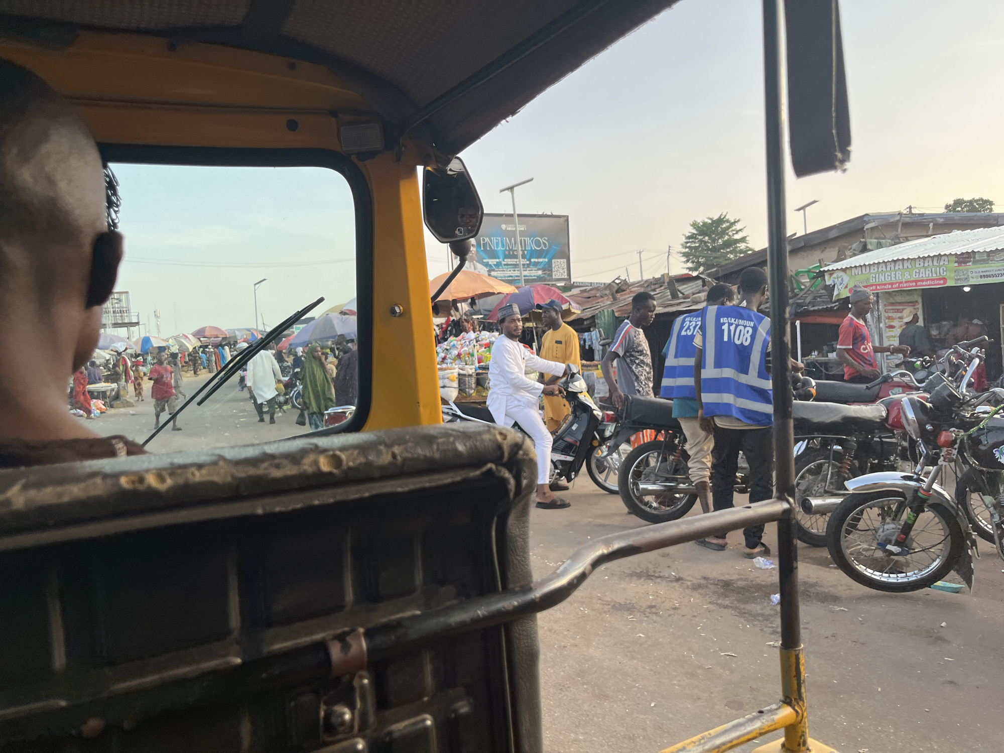 The image is taken from the interior of a yellow auto-rickshaw, looking forward through the windshield at a bustling street scene. In the foreground, the back of the driver's head and neck are visible. The street is filled with people and motorcycles. Several individuals are standing or walking, some wearing traditional clothing and others in more modern attire. Two men are wearing bright blue safety vests, one with the number "237" and the other with "1108". A man in a white outfit is seated on a motorcycle, facing forward. In the background, there are market stalls with awnings and a billboard advertising "PNEUMATIKOS". The environment appears to be a market or busy street in a city, with a somewhat dusty and unpaved road. The sky is bright, suggesting it is daytime, likely late afternoon given the lighting. The overall impression is one of a vibrant and active urban environment.