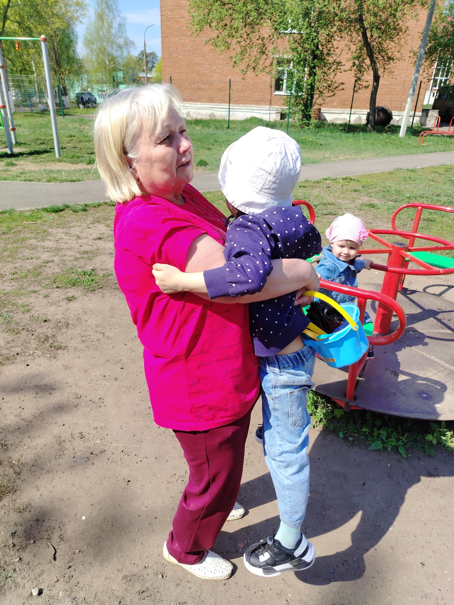 An outdoor scene in Noviy, Russia, shows an older Caucasian woman with short blonde hair wearing a bright pink top and burgundy pants, embracing a small child wearing a white hat, blue polka-dot shirt, and jeans. The child holds a blue bucket with yellow handle. To the right, a toddler in a pink hat and denim jacket stands on a red and green merry-go-round. The ground is bare dirt. In the background is a red brick building with white-framed windows and green trees. A playground is present with various equipment. The sky is blue with scattered clouds.