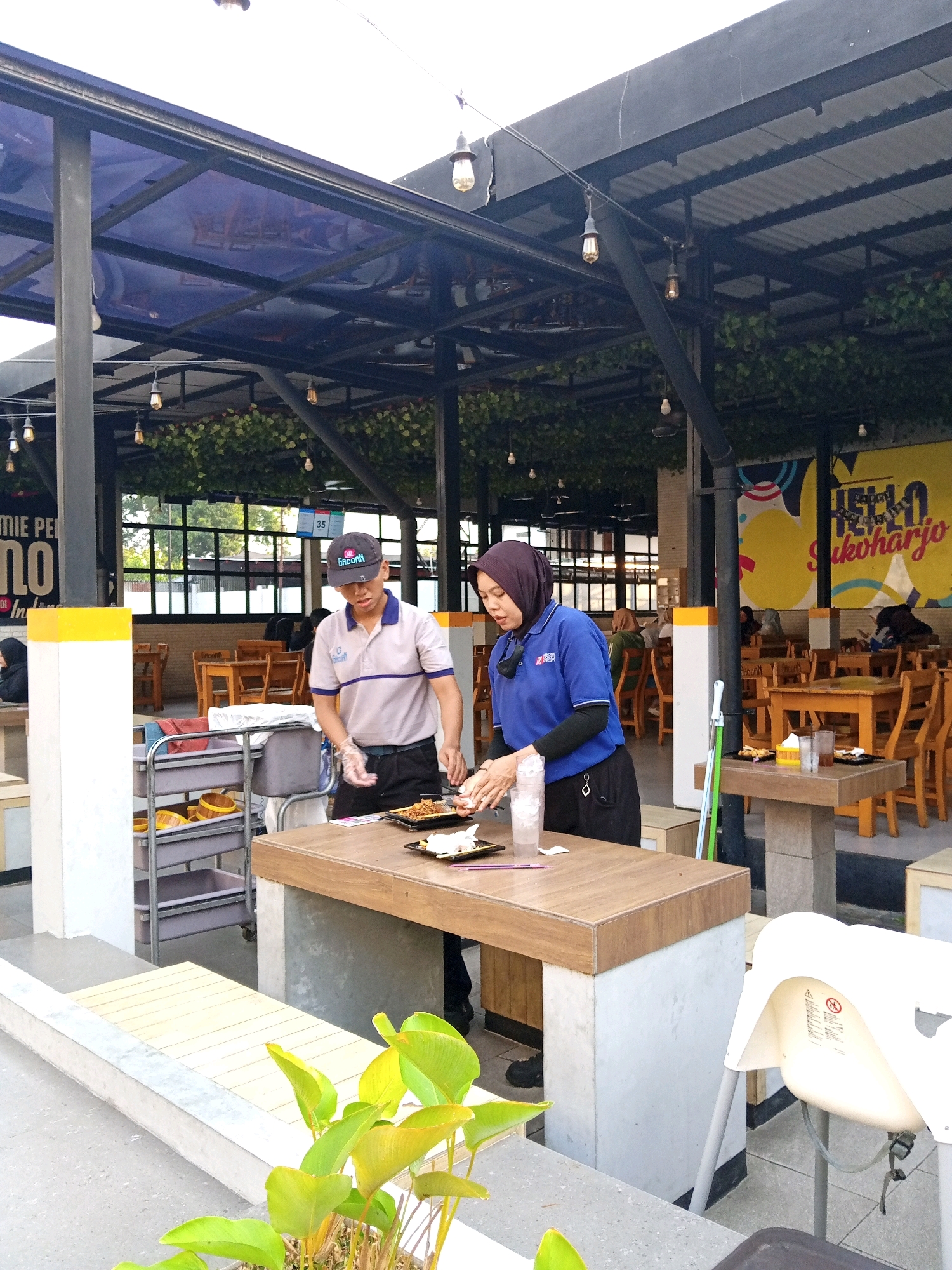 The image depicts an outdoor dining area of a restaurant, possibly in Gayam, Indonesia, characterized by its open-air structure with a corrugated metal roof and a translucent blue canopy overhead. Wooden tables and chairs are visible throughout the seating area, some adorned with potted plants and artificial ivy. In the foreground, two restaurant employees are engaged in food preparation. One, wearing a light-colored polo shirt and a dark cap, is meticulously arranging food on a plate. The other, in a blue uniform and headscarf, is also working with food and drinks. A metal serving cart with various containers stands nearby, and a white high chair is positioned at a table. A prominent yellow and white pillar, possibly a structural element or signage, is present on the left side of the frame. The background features a large yellow mural with text, and through the restaurant's open sides, glimpses of trees and windows can be seen, suggesting an urban or semi-urban setting. The overall atmosphere is casual and functional, with overhead string lights adding to the ambiance.