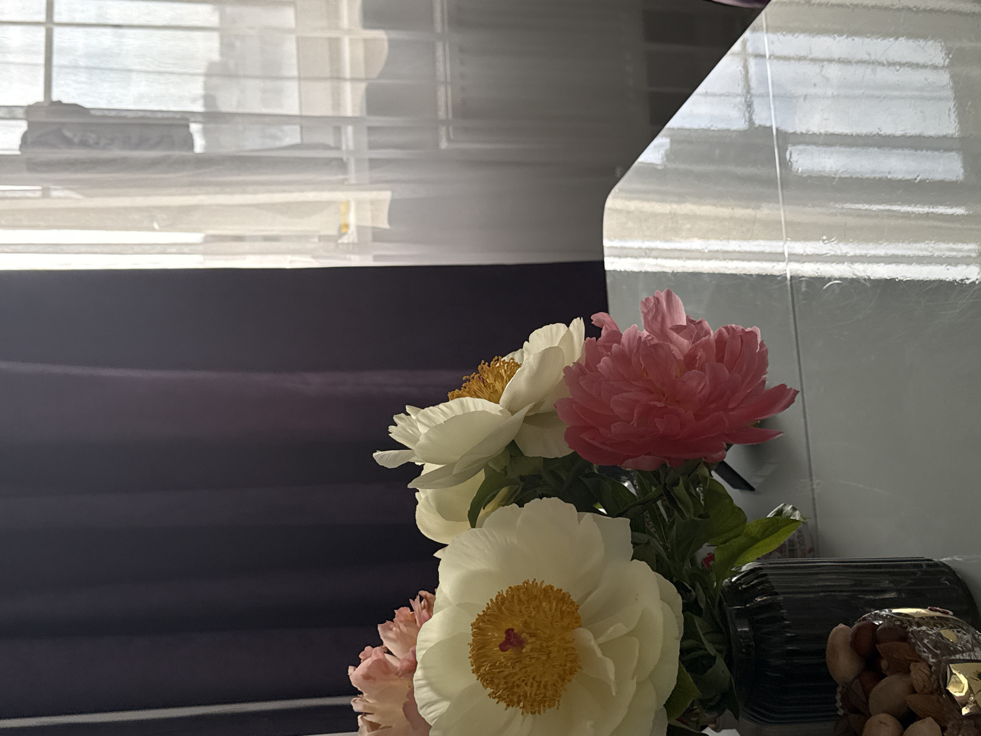 A close-up indoor shot shows a bouquet of peonies in front of a window. The flowers are a mix of white and pink, with prominent yellow centers. The window behind the flowers has white blinds, and part of a white, glossy surface (possibly a wall or cabinet) is visible on the right. A dark-colored vase partially filled with nuts is located to the right of the flowers. The environment appears to be inside a home or similar setting, but there are no people visible in the scene. The location in the city is specified as Unknown, Unknown.