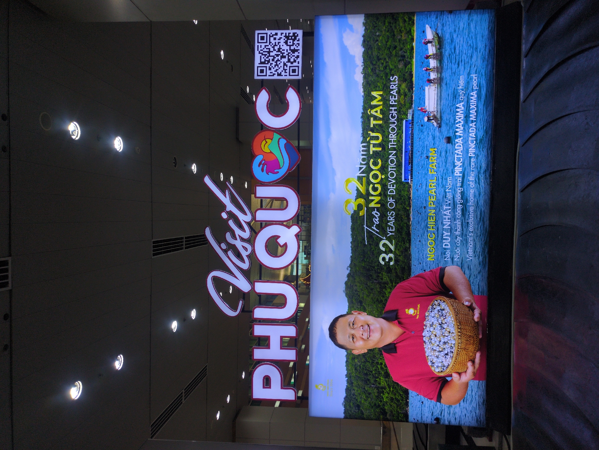The image shows a promotional sign for Phú Quốc, a city in Vietnam, focusing on pearl farming. The sign reads "Visit Phu Quoc" and features a QR code, followed by "32 Years of Devotion Through Pearls." It includes a photograph of a man holding a basket full of pearls, with a landscape of water and trees in the background. The text also identifies "Ngoc Hien Pearl Farm," calling it Vietnam's exclusive home of the rare Pinctada Maxima pearl. The sign is illuminated, suggesting it's likely located in a public space, possibly an airport or tourist information center.