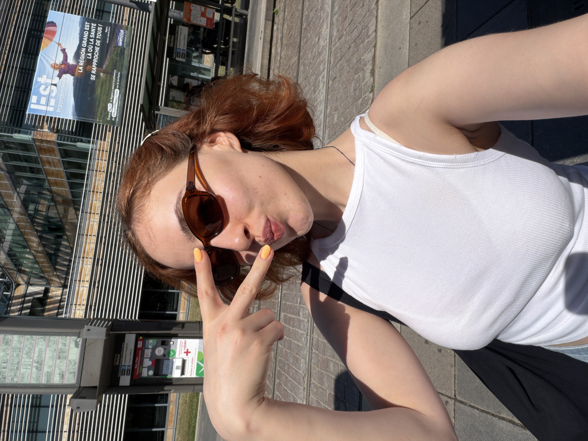 The image captures a young woman with reddish-brown hair, wearing sunglasses and a white tank top, posing for a selfie outdoors. She is making a peace sign with her right hand and puckering her lips. In the background, a modern building with large windows and a billboard advertising "LA RÉGION GRAND EST" is visible. A ticket vending machine and tram tracks are also present, suggesting an urban setting. The presence of tram tracks and the advertising billboard point to the location being in Strasbourg, France, specifically near a tram stop. The lighting indicates it is a sunny day.