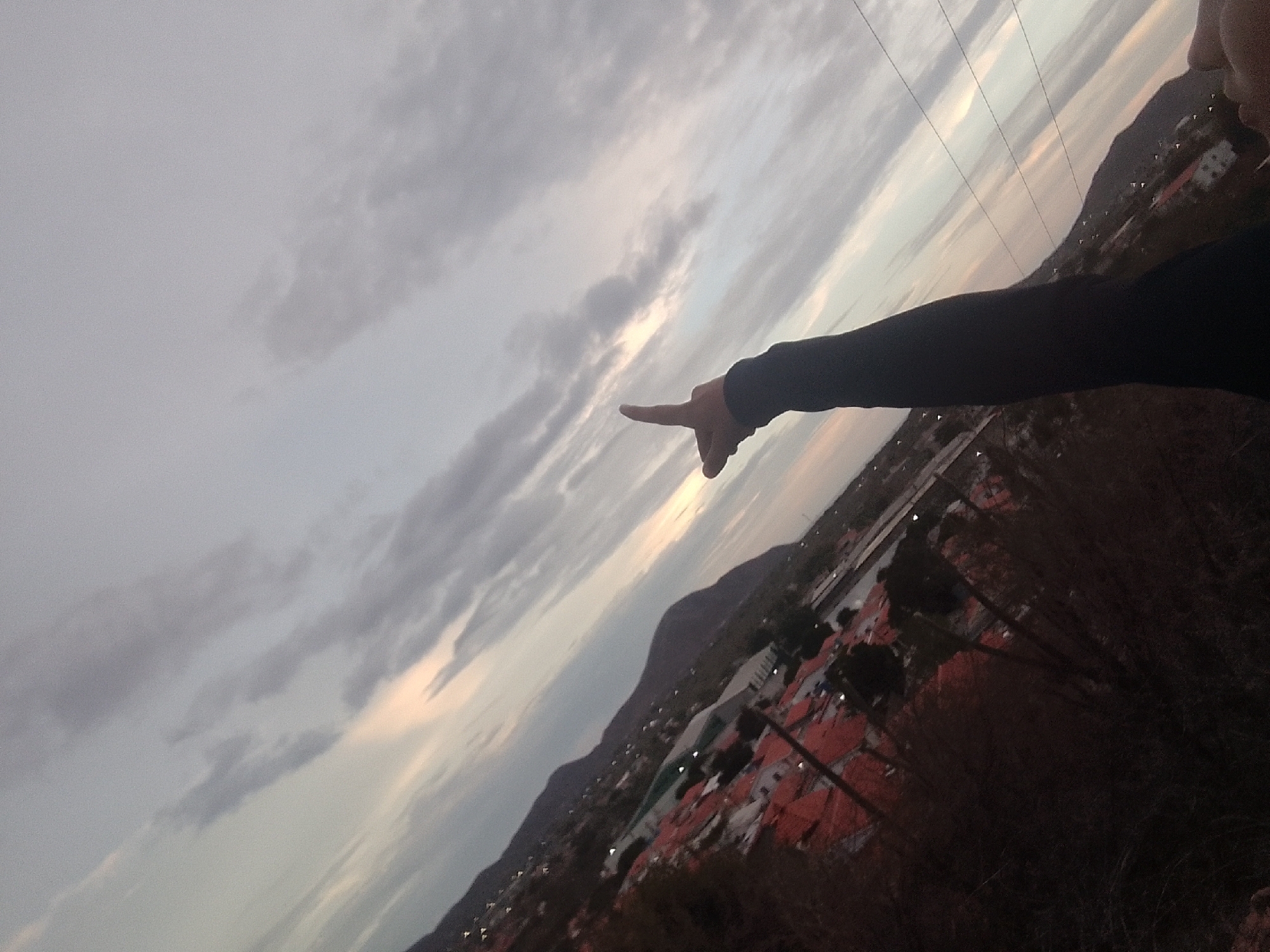 A tilted, wide-angle shot captures a view from an elevated position, likely overlooking Juan Griego, Venezuela. A person's arm, clad in a black, long-sleeved garment, extends into the frame from the upper right, with the index finger pointing toward the sky.
The sky is filled with clouds and soft light. Below, the landscape reveals a combination of residential buildings with red rooftops, interspersed with trees. Hills or mountains form the horizon line. Power lines stretch across the upper right portion of the frame. The image has a slight graininess, possibly due to low light conditions or camera settings.