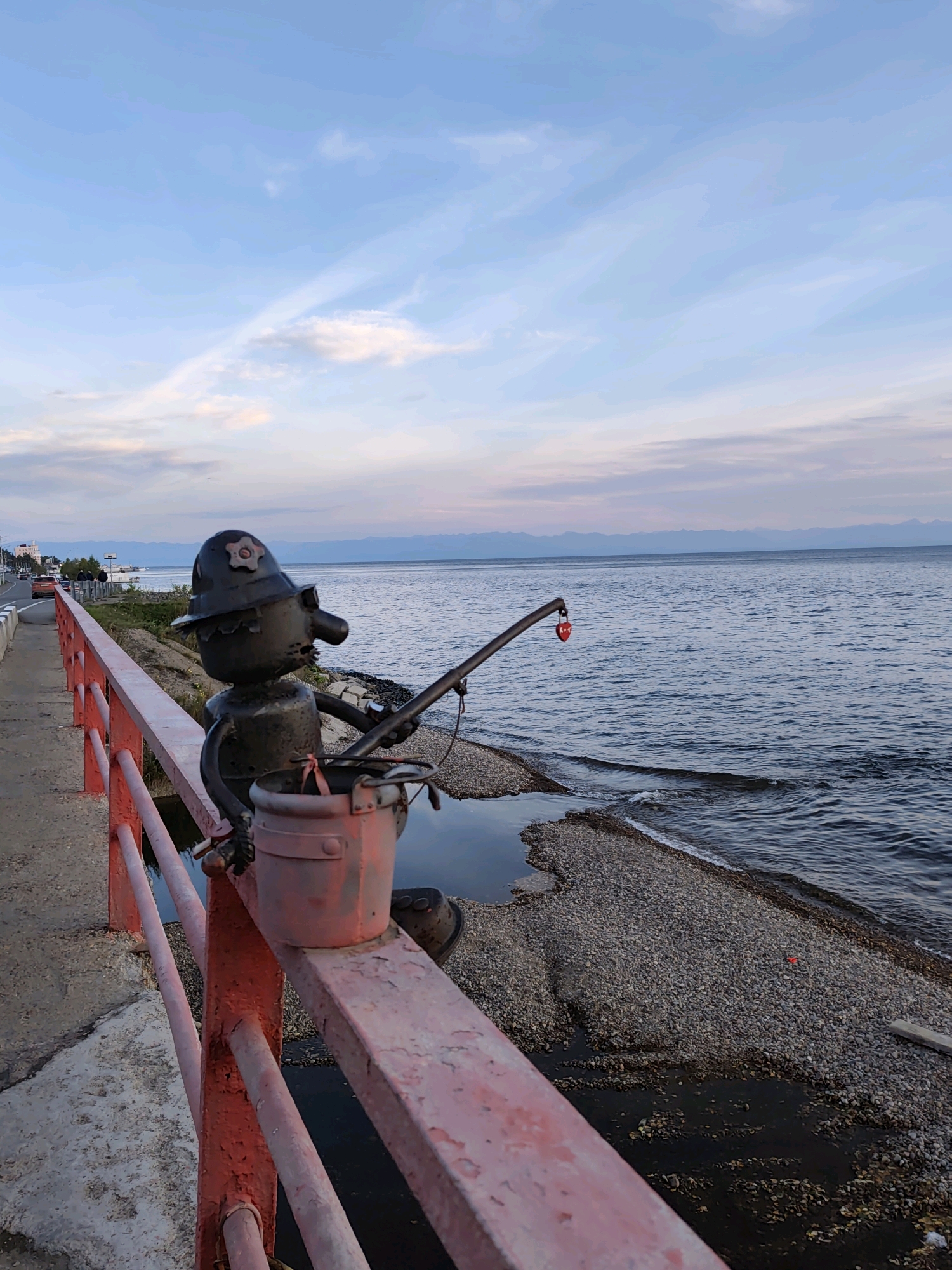 A metal sculpture of a fisherman with a bucket and fishing rod stands on a red railing overlooking a body of water. The fisherman sculpture is black, with a helmet and a red heart-shaped lock hanging from the fishing rod. The railing is weathered and red. A cobblestone beach lines the edge of the water, with small waves lapping at the shore. In the background, a distant mountain range is visible under a pale blue sky with wispy clouds. A road with cars can be seen to the left of the scene, suggesting it is located in a populated area, likely in the city of Listvyanka, Russia, known for its proximity to Lake Baikal.