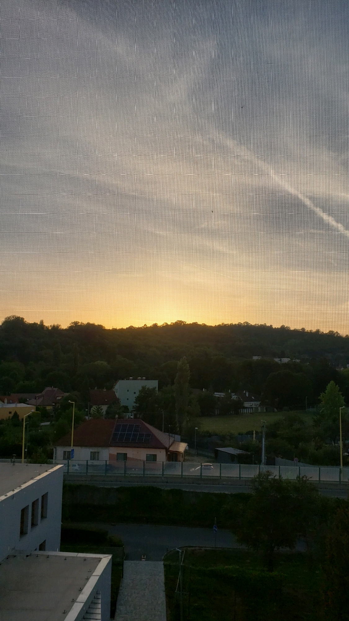 The image depicts a view from a window with a screen, looking out at a landscape at dusk. A soft, orange glow indicates the setting sun behind a tree-covered hill. In the foreground, there is a modern building with multiple windows. Further back, a house with solar panels on its roof is visible, along with other residential buildings nestled among trees. A road runs horizontally across the mid-ground, with a barrier and some streetlights. The sky is a gradient of pale blue to orange. The scene suggests a residential or semi-urban area, possibly on the outskirts of the city, with a mix of nature and man-made structures.