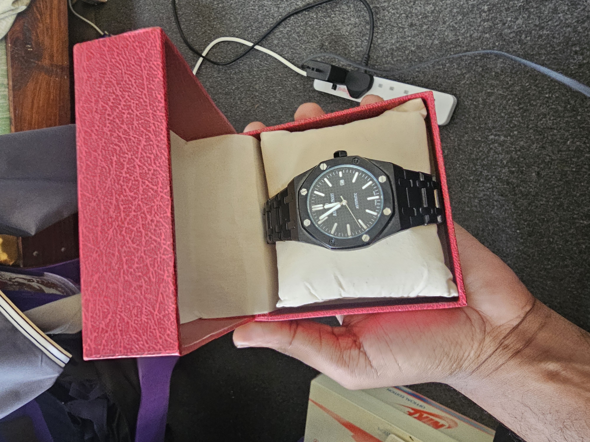 In Dar es Salaam, Tanzania, a person's hand holds a black Audemars Piguet watch within a textured pink gift box. The watch features a dark dial with silver hour markers and hands, and the word "AUTOMATIC" is visible at the bottom. The box is open, revealing the watch resting on a cream-colored cushion. In the background, a white box with red accents, possibly containing Nike shoes, is partially visible, along with electrical cords and charging devices on a grey carpeted surface.
