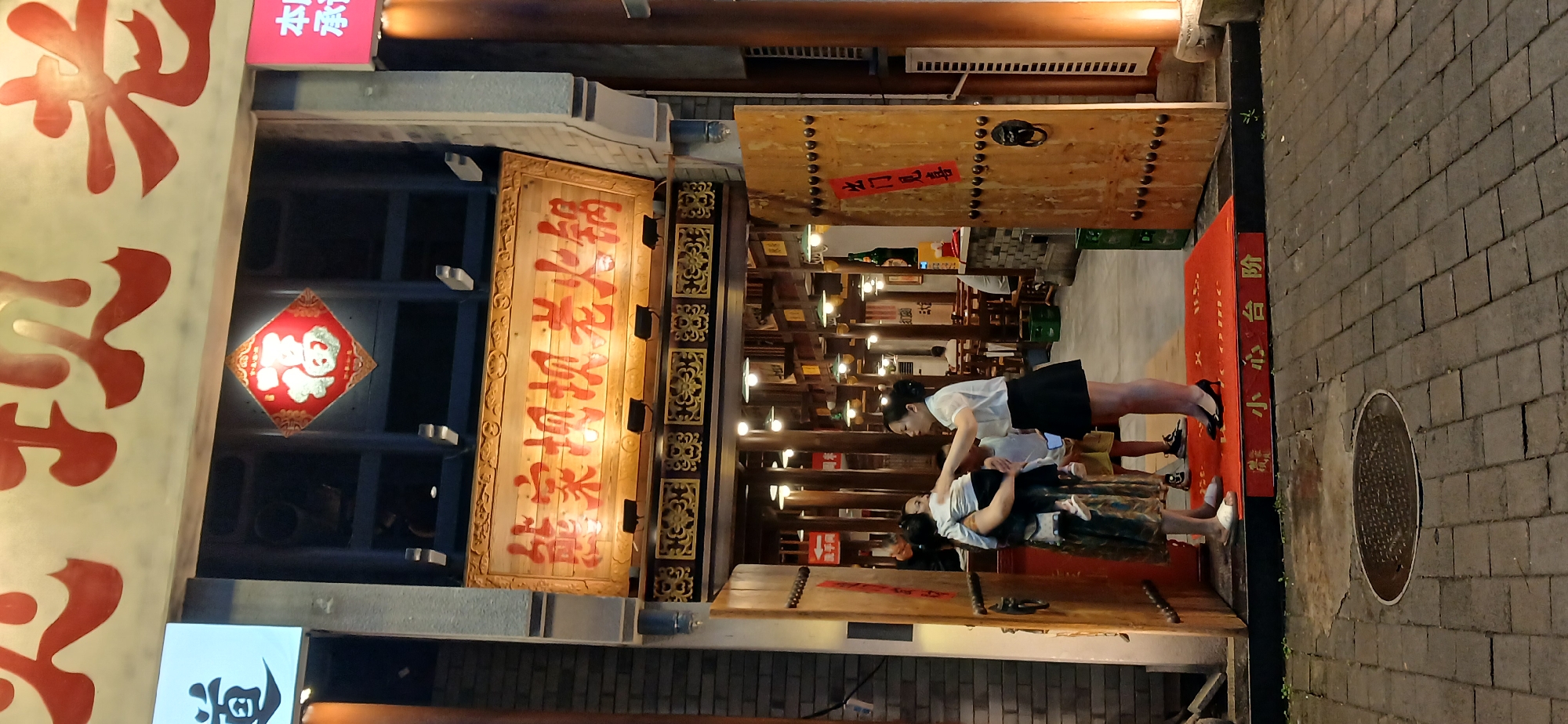 A vertical, outdoor, eye-level shot captures the entrance to a restaurant named "老火锅" (Lao Huo Guo), or "Old Hot Pot" in English, located in the Jiulongpo District, China. The establishment features traditional wooden doors and signage, adorned with red Chinese characters and decorative elements. A red welcome mat with gold characters reading "小心台阶" (Xiǎoxīn táijiē), meaning "Beware of steps," is laid out on the cobblestone pavement.
In the foreground, a woman in a patterned dress and sandals holds a young child. Beside her, a woman in a white short-sleeved shirt and black skirt stands, appearing to interact with the child. Another child, wearing a yellow shirt and shorts, stands behind the woman in the dress. The entrance is illuminated by warm interior lighting and decorative lamps hanging from the ceiling. A large red square ornament with gold Chinese characters, likely representing good fortune, is mounted above the entrance. The overall scene conveys a welcoming atmosphere typical of a traditional Chinese eatery.