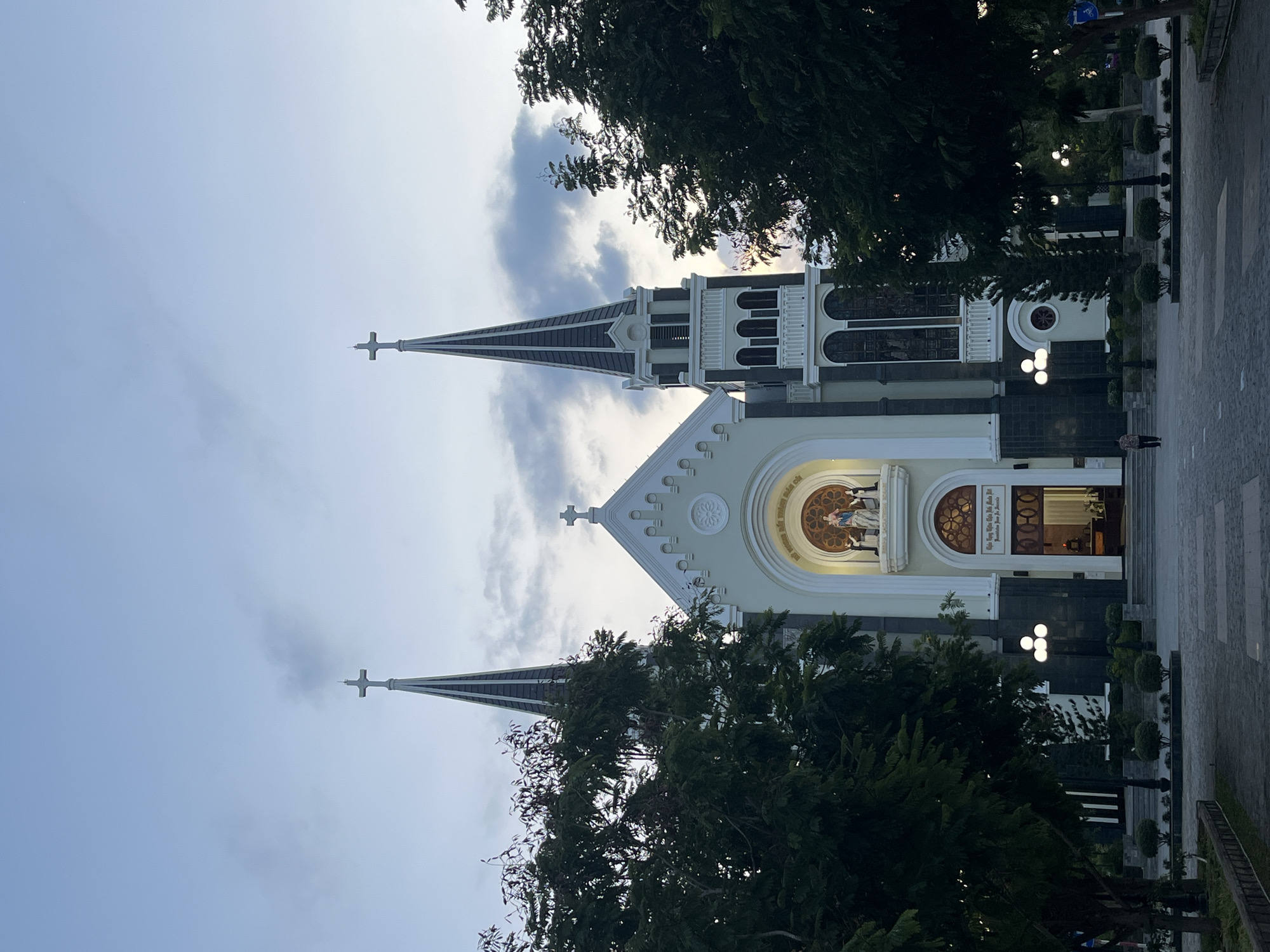 The image shows the exterior of a church, identified as the Sacred Heart Cathedral, located in Bac Nha Trang Ward, Vietnam. The cathedral features two prominent spires topped with crosses, flanking a central facade with arched windows and a large circular stained-glass window above the entrance. A statue of Mary holding the baby Jesus is visible in a niche above the entrance, flanked by two kneeling figures. The church is surrounded by lush green trees and a paved courtyard with a few streetlights and small, manicured bushes. The sky is overcast with a hint of twilight. A single person is walking away from the camera in the courtyard, dressed in a patterned shirt and dark pants. The overall scene depicts a quiet, late afternoon or early evening setting outside a significant religious building.