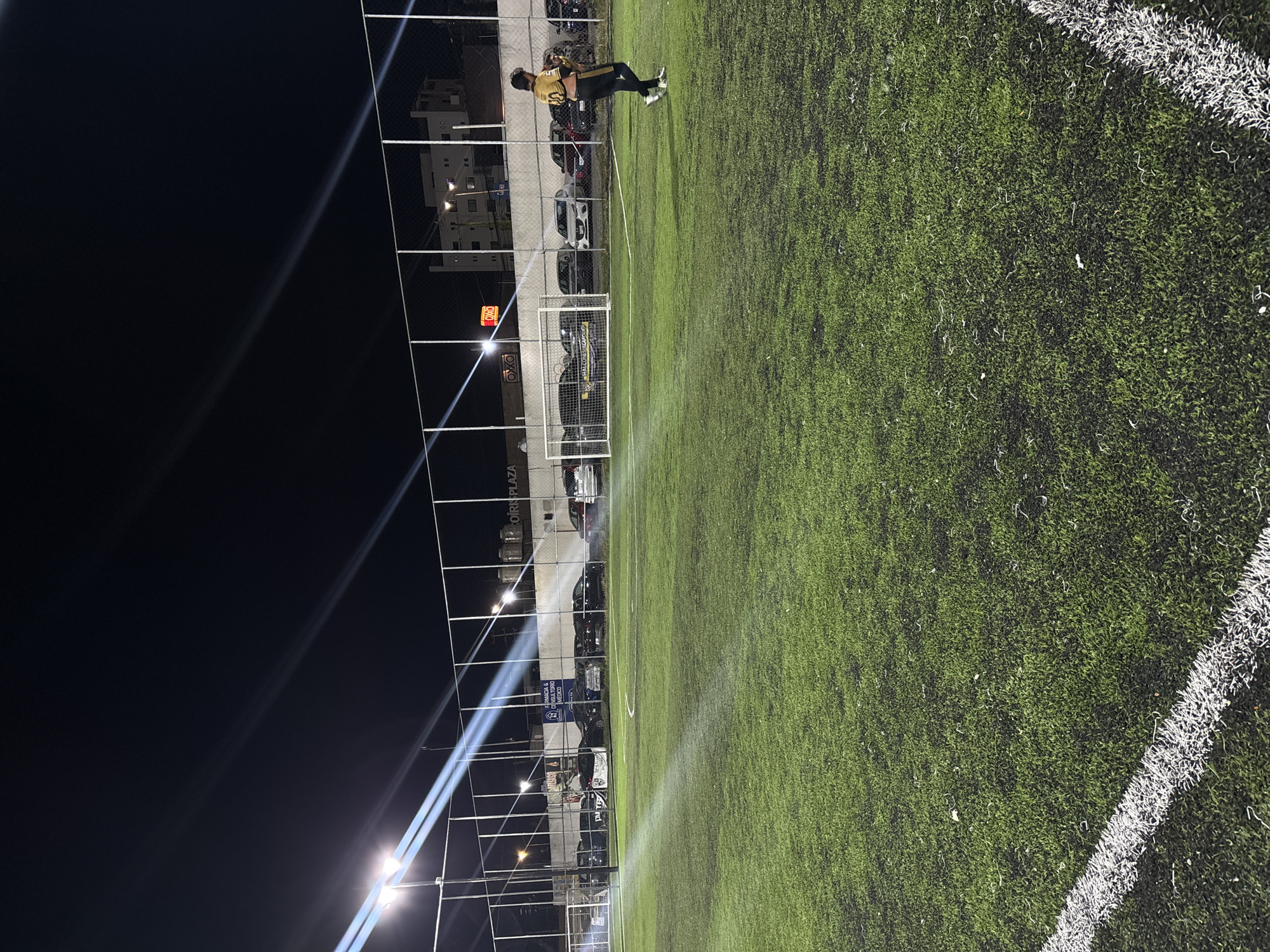 The image shows a brightly lit outdoor soccer field with artificial turf at night in San Bernardino Tlaxcalancingo, Mexico. A person in a black and gold soccer uniform is captured in motion on the field. Adjacent to the field is a chain-link fence, behind which are parked cars and buildings. A street sign for "OIR PLAZA" is visible behind the fence, and there are additional street signs, one for "FARMACIA & CONSULTORIO MEDICO INVERSIONES." Bright stadium lights illuminate the scene, casting strong beams of light into the dark sky. The field's white boundary lines are clearly visible against the green turf. A soccer goal is positioned behind the person, and a commercial establishment is also visible behind the fence.