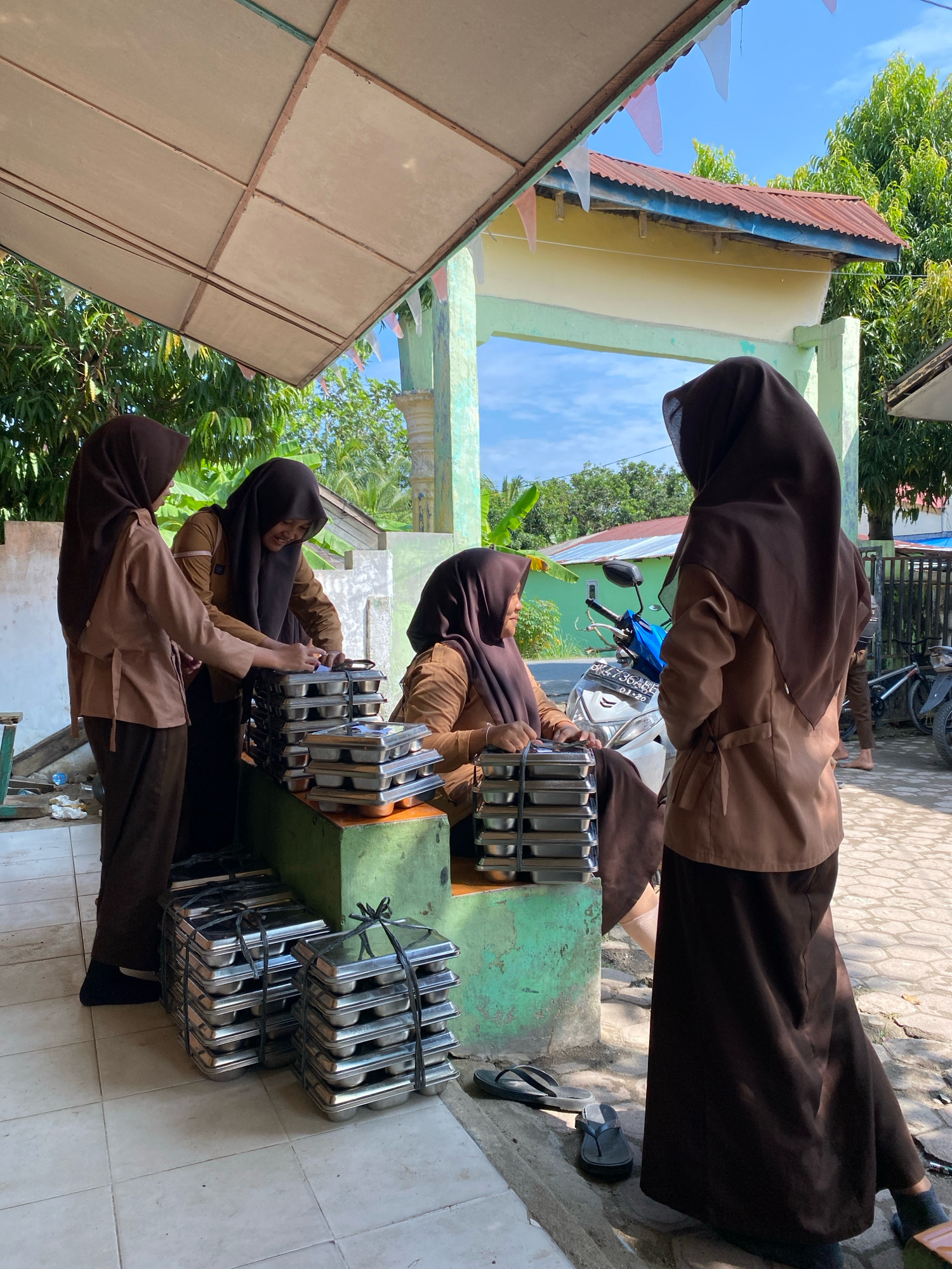 Four individuals, likely students, are gathered outdoors in what appears to be a school or communal area in Karang Gading, Indonesia. They are all wearing brown uniforms and headscarves. Two of the individuals are standing near a green-painted structure, interacting with stacks of metal food trays. One person is seen counting or organizing the trays, while another is looking on. Two other individuals are standing further back; one is facing away from the camera, and the other is partially obscured.
In the background, there is a building with a yellow facade and a red corrugated metal roof. To the right, a fence is visible, and further back, trees and a blue sky suggest a sunny day. A blue and white motorcycle is parked behind the individuals. The ground is a tiled patio. The scene suggests a food distribution or preparation activity.