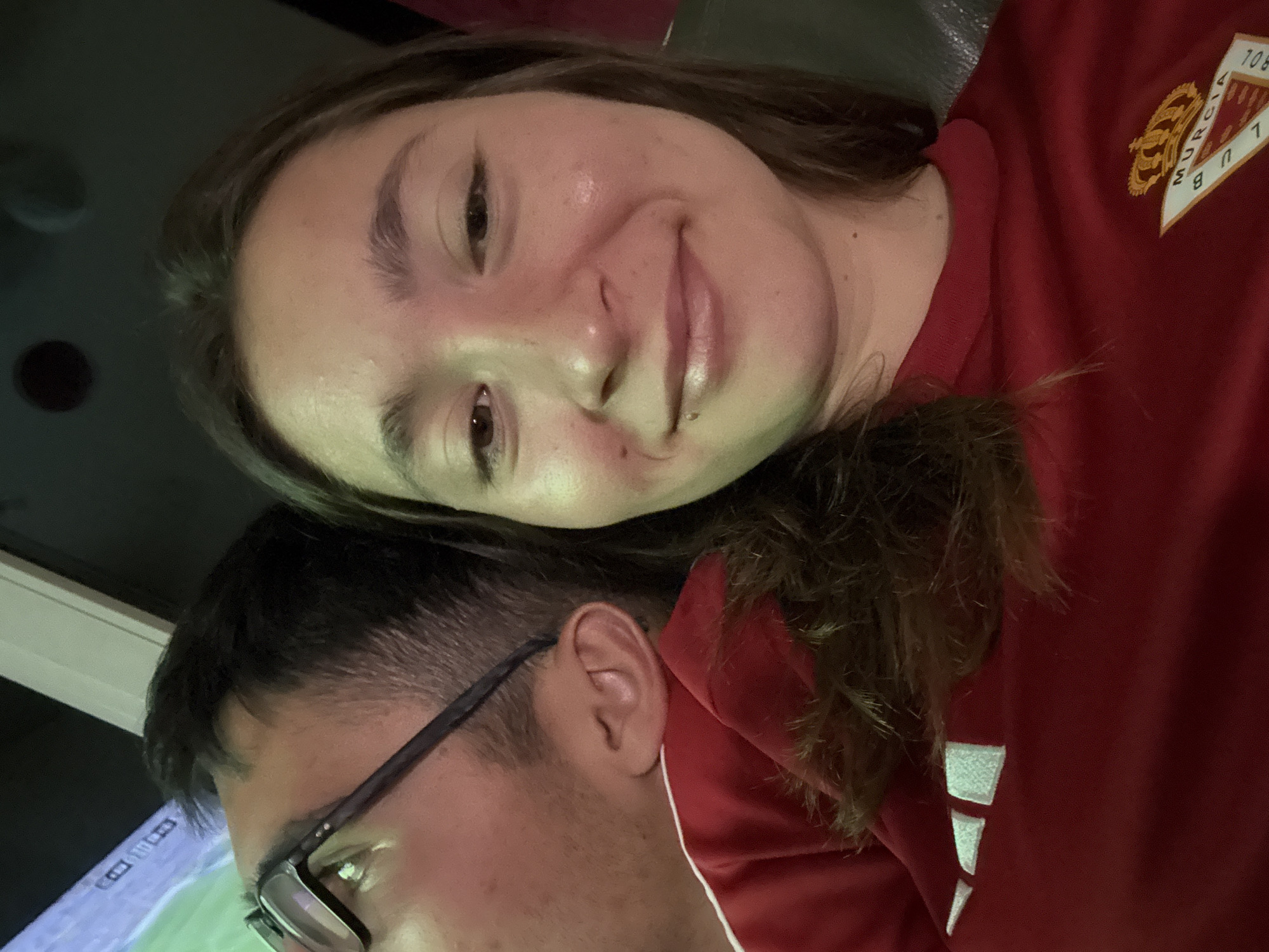 Two individuals, a male and a female, are positioned closely together, with the male's head leaning on the female's shoulder. The male is wearing glasses and a red shirt with white accents. The female is also wearing a red shirt, with a crest on it that reads "Murcia F.C.". The setting appears to be indoors, possibly a living room or a place with a large screen displaying a video game, indicated by the green-hued lighting on the male's face and the visual content on the screen. The lighting suggests it is nighttime or a dimly lit environment. The overall atmosphere is casual and intimate. The image does not provide any specific visual cues to place the location in Sofia, Bulgaria.