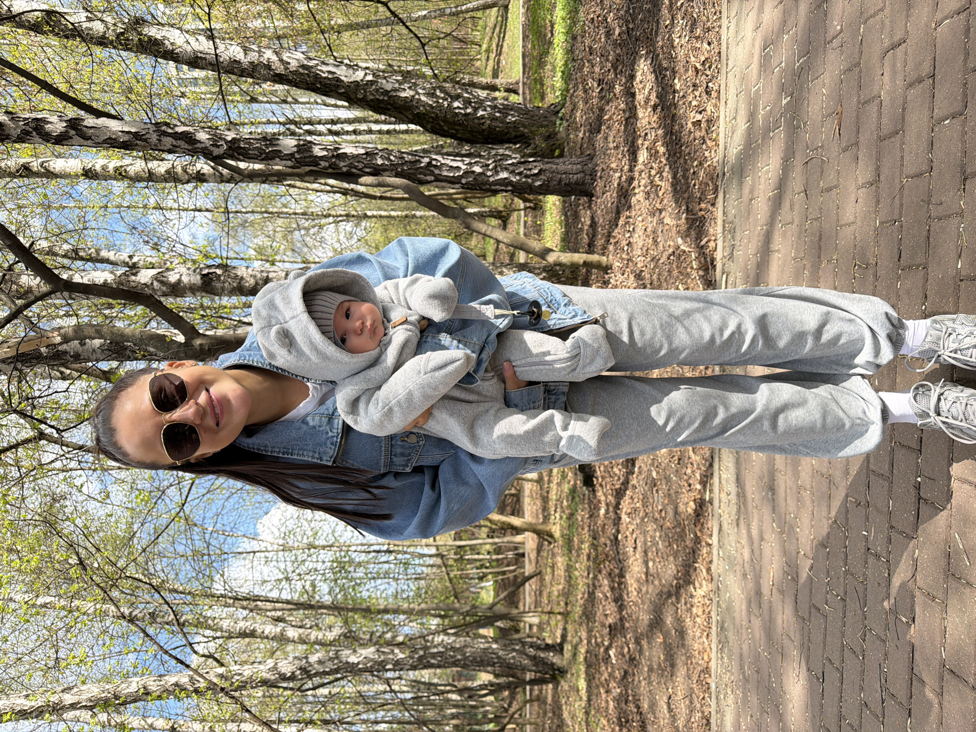 A woman stands on a brick-paved path, holding a baby, likely in Khimki, Russia, based on the presence of birch trees, which are common in that region. The woman wears sunglasses, a denim jacket, grey sweatpants, and white socks with grey sneakers. She has long, dark hair. The baby is dressed in a grey hooded outfit with a matching hat. The background includes bare trees with budding leaves and leaf-covered ground.