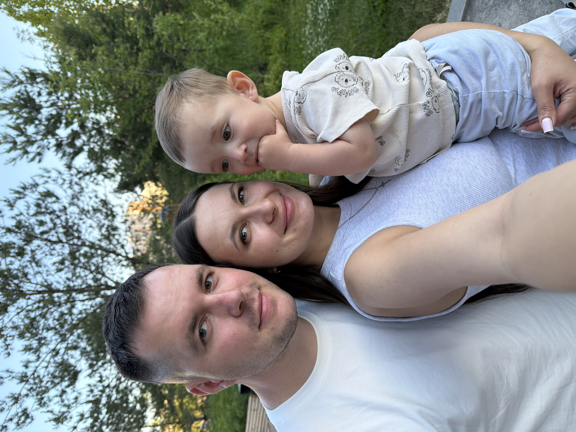 A portrait-oriented photograph depicts three individuals outdoors. On the left, an adult male with short dark hair and light stubble, wearing a white t-shirt, is positioned next to an adult female. He faces the camera directly. The adult female, positioned centrally, has long dark hair and wears a light gray ribbed tank top with a silver necklace featuring a small cross pendant. She holds an infant/toddler in her left arm and also faces the camera directly. The infant/toddler, with light-colored hair, wears a light beige short-sleeved hooded top patterned with cartoon lions and light blue pants. The child is looking towards the right side of the frame, with its right hand raised to its mouth. The background consists of blurred green foliage and trees beneath a clear blue sky, indicating a natural outdoor environment, possibly a park. A light-colored wooden structure is partially visible behind the adult male. The lighting is natural and even. The specific location of Moscow, Russia, cannot be identified from the visual information present in the image.