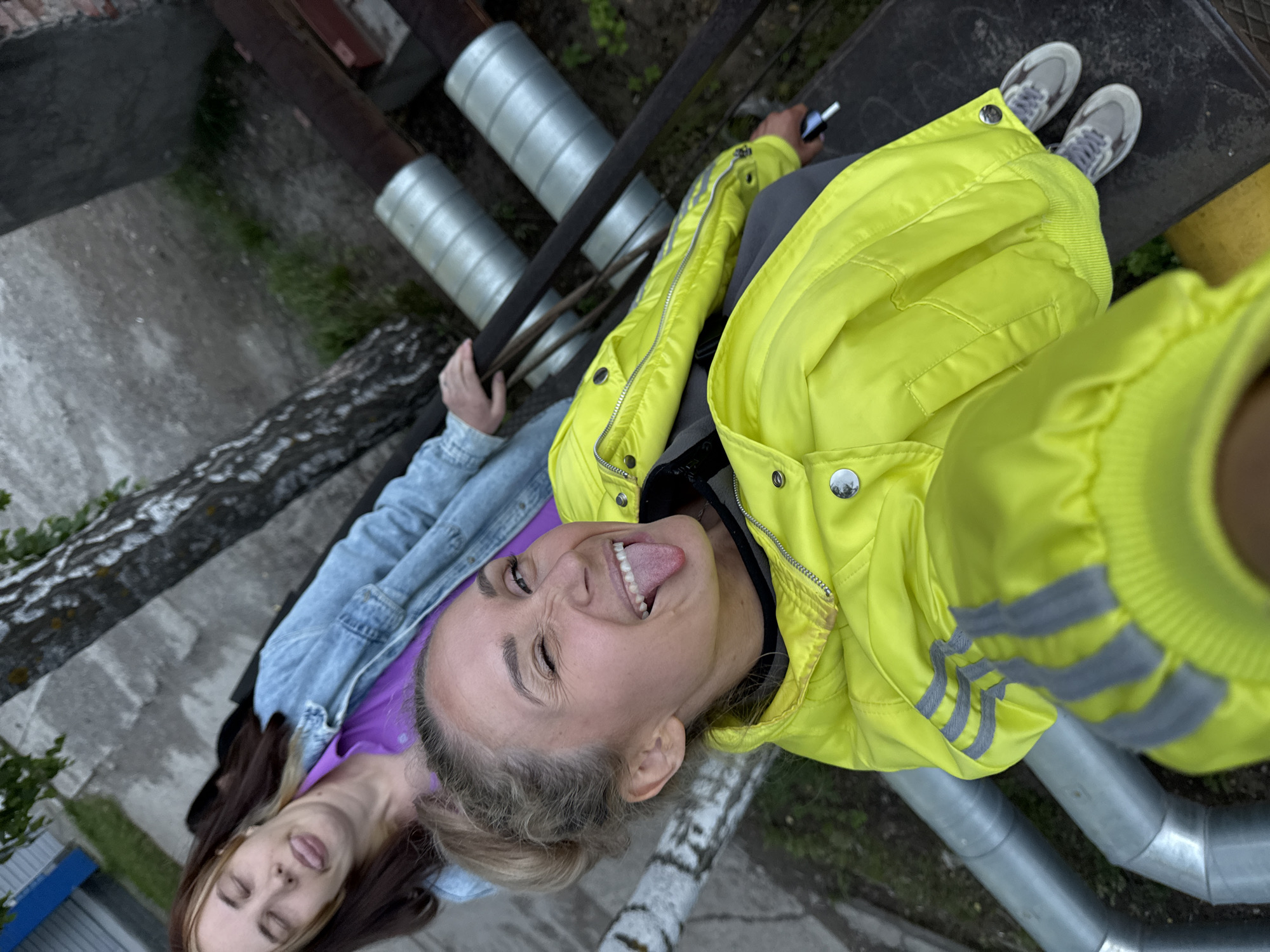 The image depicts an outdoor scene in the city of Chaikovsky, Russia, featuring two women in a playful interaction. In the foreground, a woman with blonde hair tied in a bun winks her left eye and extends her tongue with a wide, open-mouthed grin. She wears a bright neon yellow jacket with grey reflective stripes on the shoulders, a dark undershirt, dark pants, and grey sneakers. Her left hand holds a small black electronic device, possibly a vape pen. She appears to be seated on a dark, flat surface.
Behind her, another woman with long dark brown hair and lighter streaks poses with her eyes closed and tongue also extended. She is dressed in a light blue denim jacket over a vibrant purple collared shirt. Her right hand rests on a dark, horizontal metal beam or railing positioned between her and the foreground woman.
The background reveals a series of insulated silver-colored pipes running horizontally across the frame, likely part of an industrial or utility system. To the left, a birch tree with distinctive white and black bark and sparse green leaves is visible. The ground consists of a dark concrete or paved area that transitions into dirt and sparse green vegetation. Further in the background, a grey concrete wall and a hint of a blue structure are discernible under an overcast sky, indicating an urban or semi-industrial setting.