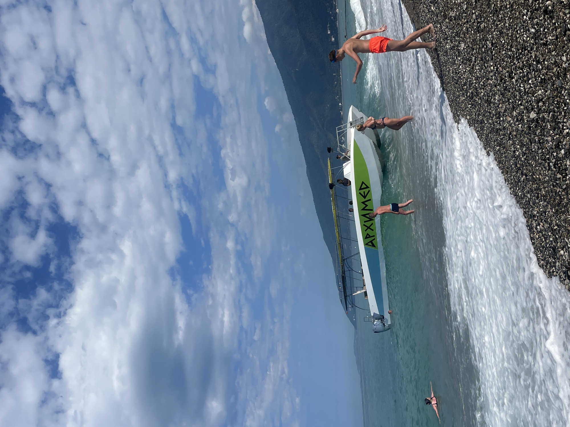 The image depicts a coastal scene in Gagra, Abkhazia, with a catamaran boat moored near the shore. The sea is a vibrant turquoise, with gentle waves lapping against a pebbled beach. A lush green mountain range forms a scenic backdrop to the scene, its slopes dotted with trees. The sky is a dynamic mix of blue and white clouds, suggesting a pleasant, partly sunny day.
In the foreground, several people are enjoying the beach. A young boy in blue swim trunks stands in the shallow water, his back to the camera, facing the boat. To his right, a woman in a black bikini wades in the water, her gaze directed towards the boat. Further to the right, a young man in bright orange swim trunks stands on the pebbled shore, his left arm extended as if pointing or gesturing. His gaze is also directed towards the boat.
The catamaran itself is white with a green stripe and bears Cyrillic lettering that reads "АРХИМЕД" (Arkhimed). It appears to be a passenger boat, with several individuals visible on its deck, some standing and others seated. The boat is partially submerged in the water, indicating it is either at anchor or in the process of docking.
On the left side of the frame, a person with dark hair is swimming in the sea, lying on their back and looking up at the sky.
The overall atmosphere is one of leisure and recreation, capturing a moment of activity at the seaside in Gagra.