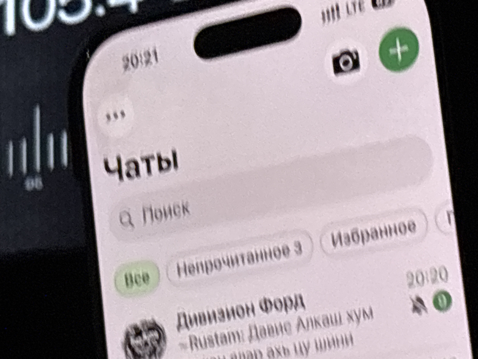 This is a close-up, high-angle shot of a smartphone screen displaying a messaging application. The screen shows a list of chats, with the title "Чаты" (Chats) prominently displayed. The time "20:21" is visible in the top left corner of the screen. Below the title, there is a search bar with a magnifying glass icon and the text "Поиск" (Search).
Further down, there are several chat categories or filters, including "Все" (All), "Непрочитанное 3" (Unread 3), and "Избранное" (Favorites). A chat entry shows a profile picture with a person's face, followed by "Дивизион форд" (Division Ford) and a message starting with "-Rustam: Давнс Алказ хүм". The time "20:20" is also visible next to this chat entry, along with icons indicating no sound and a notification.
The phone itself appears to be a modern smartphone with a notch at the top, common in devices like iPhones. The overall environment is dark, suggesting the photograph was taken in low light conditions, possibly indoors. There are no identifiable landmarks or elements that would allow for the specific location in Grozny, Russia, to be determined. The image focuses solely on the content displayed on the phone screen.
