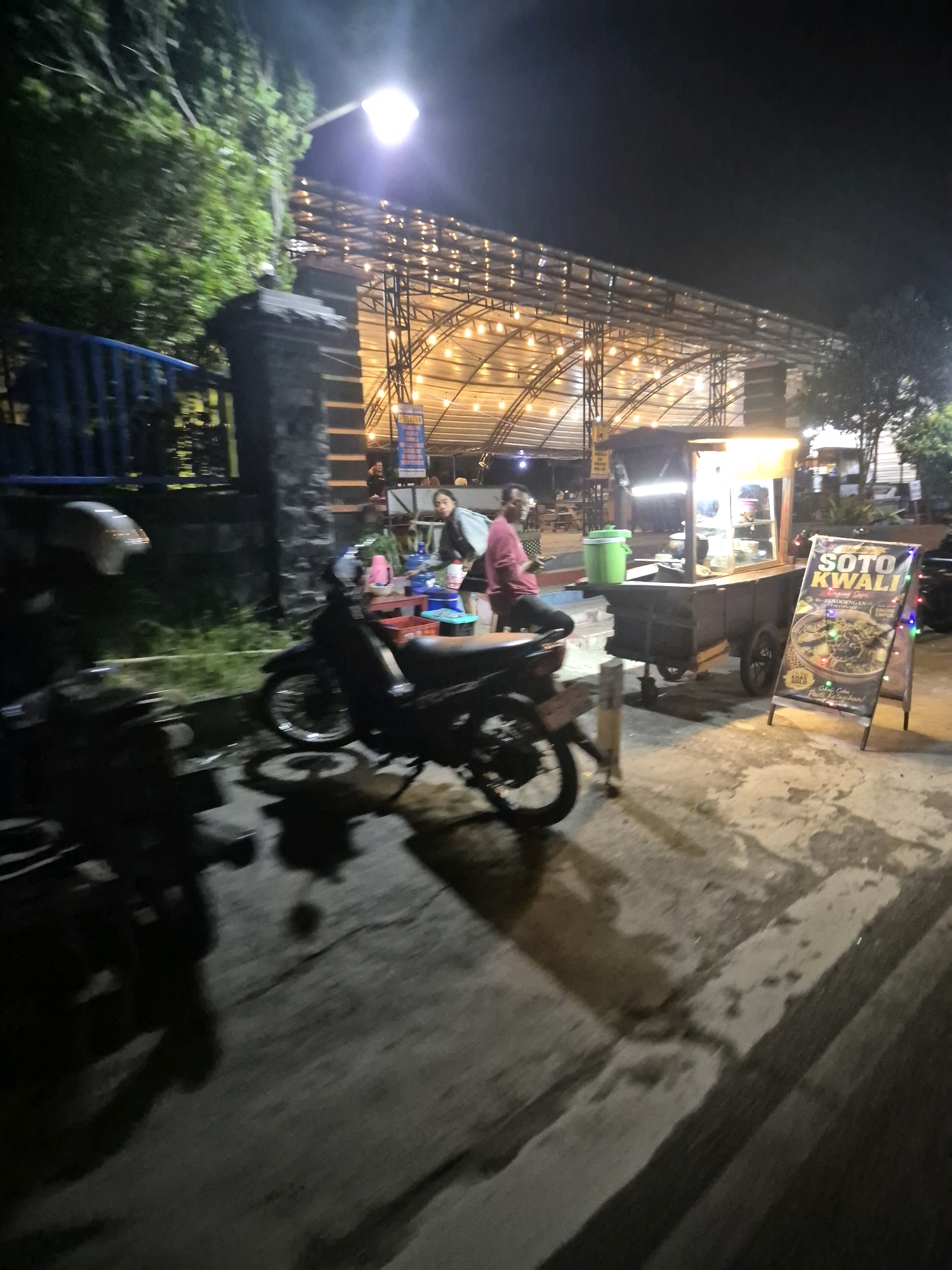 This is a night time photograph taken in the city of Surakarta, Indonesia, showing an outdoor food stall. The stall is a cart with a glass display case for food, illuminated by overhead lights. A sign in front of the cart advertises "Soto Kwali" with a picture of the dish. Two people are visible near the stall; one is seated and wearing a pink shirt, while another is standing and looking towards the camera. Several motorcycles are parked in the foreground, and a covered seating area with string lights is visible in the background. The scene appears to be on a street or walkway with a concrete surface.