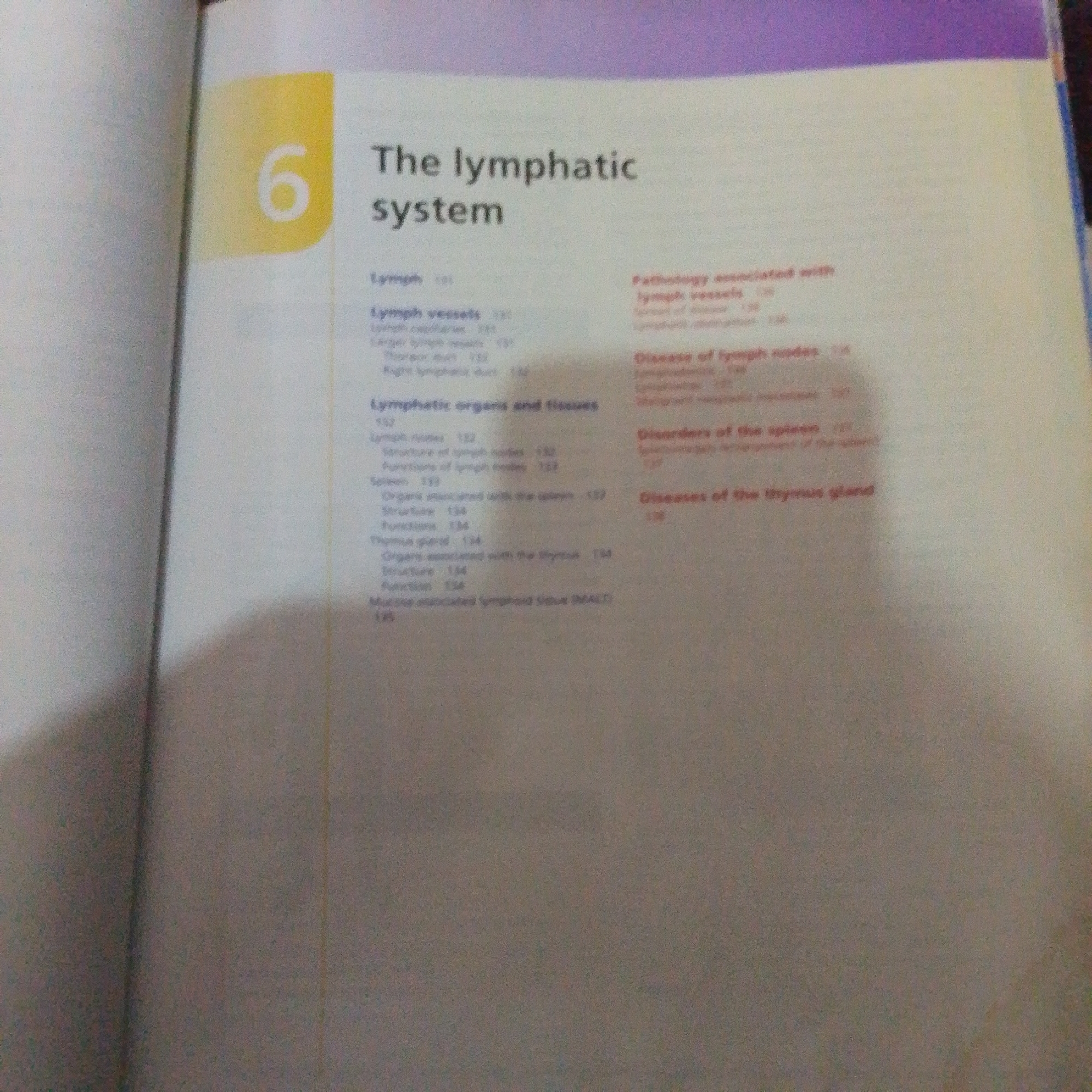 The image is a close-up photograph of an open book, likely a textbook or reference material, lying flat. The foreground is dominated by the right-hand page, which contains a table of contents or index. The left-hand page is mostly obscured by shadow and is out of focus, but a large yellow numeral "6" is visible in a colored block.
The visible text on the right page is printed in black and red. The black text includes headings like "Lymph," "Lymph vessels," and "Lymphatic organs and tissues." Under these headings, there are sub-entries such as "Lymph capillaries," "Larger lymph vessels," "Thoracic duct," "Right lymphatic duct," "Lymph nodes," "Structure of lymph nodes," "Functions of lymph nodes," "Spleen," "Organs associated with the spleen," "Structure," "Functions," "Thymus gland," "Organs associated with the Thymus," "Structure," "Function," and "Mucosa-associated lymphoid tissue (MALT)." Some of these entries are followed by page numbers, for example, "131," "132," "133," "134," and "135."
The red text appears in a column on the right side of the page, with headings like "Pathology associated with lymph vessels," "Disease of lymph nodes," and "Diseases of the thymus gland." These also have sub-entries and page numbers.
The overall setting is that of someone reading or referencing a book. The lighting suggests indoor conditions, and the image has a slightly grainy quality. There are no people or explicit environmental cues to indicate the time of day, weather, or a specific location beyond the book itself. The context provided about Jalingo, Nigeria, does not have any visual correlation within the image content.