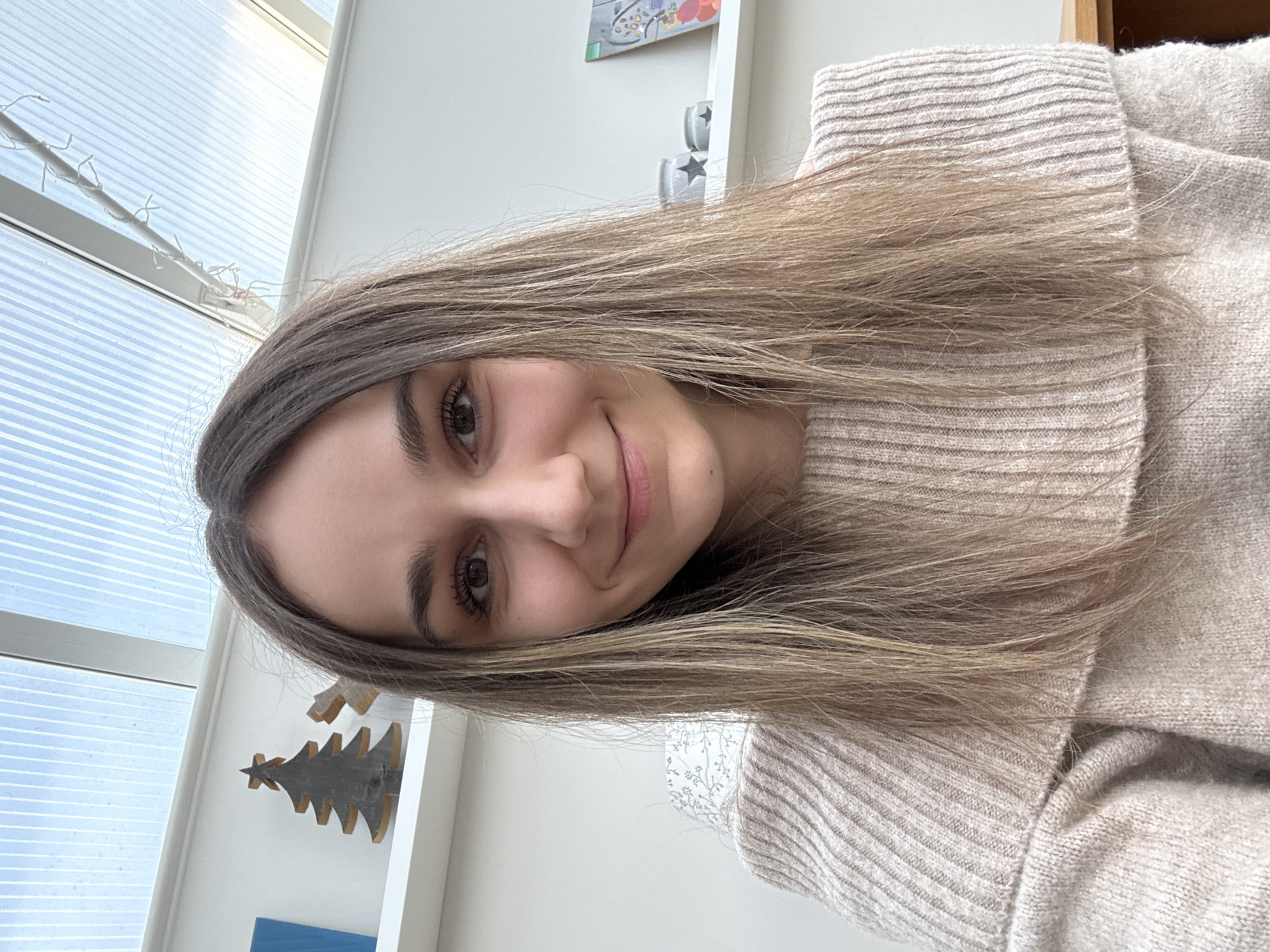 A young woman with long, layered, highlighted brown hair is shown in a close-up shot, partially rotated counter-clockwise. She is wearing a beige ribbed knit sweater with a high neckline. She is slightly smiling and looking directly at the camera.
The background includes a white shelf with decorative items like a Christmas tree figurine and star-patterned containers. A window with white frames and blinds is visible in the background. The location cannot be determined within the city of East Suffolk, United Kingdom based on visual evidence alone.