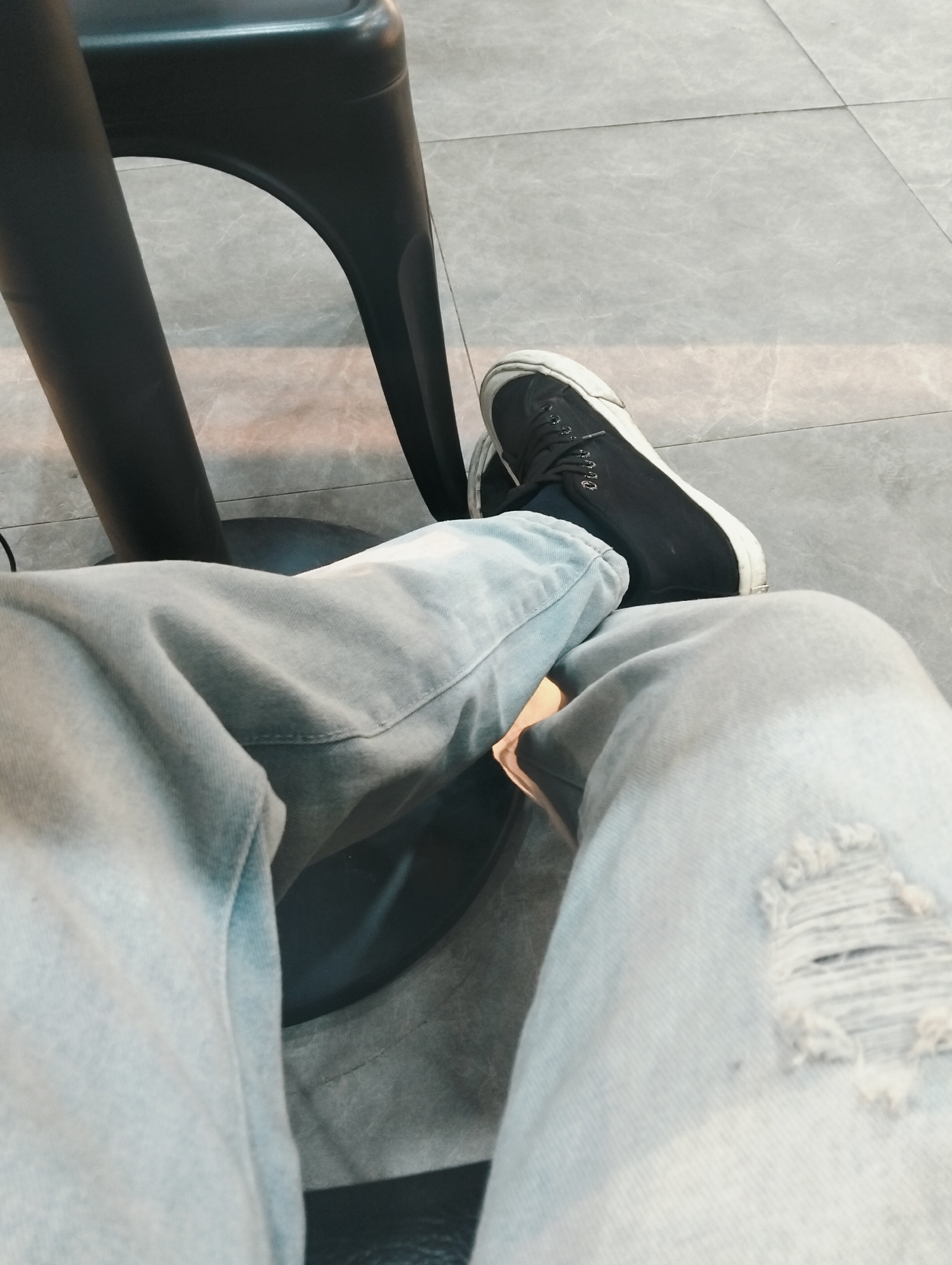 The image shows a person's legs, clad in light-wash ripped jeans, resting on a black stool or chair. The person is wearing black sneakers. The setting appears to be indoors, with a tiled floor visible. The scene is captured from a low-angle perspective, focusing on the lower body and footwear.