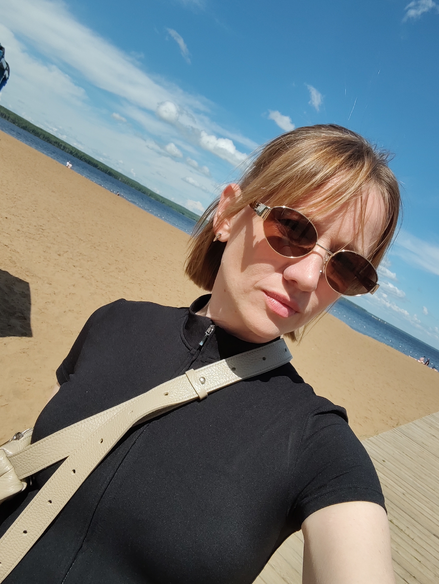 A medium shot captures a woman with shoulder-length blonde hair and sunglasses at a beach. She is wearing a black t-shirt and a light-colored crossbody bag. Her head is tilted slightly downwards, and she is looking towards the camera. The background shows a sandy beach, a body of water with a tree-lined distant shore, and a blue sky with scattered clouds. The wooden planks of a boardwalk are visible in the foreground on the right. The image appears to have been taken on a sunny day. It is likely the Kama River beach in Chaikovsky, Russia.