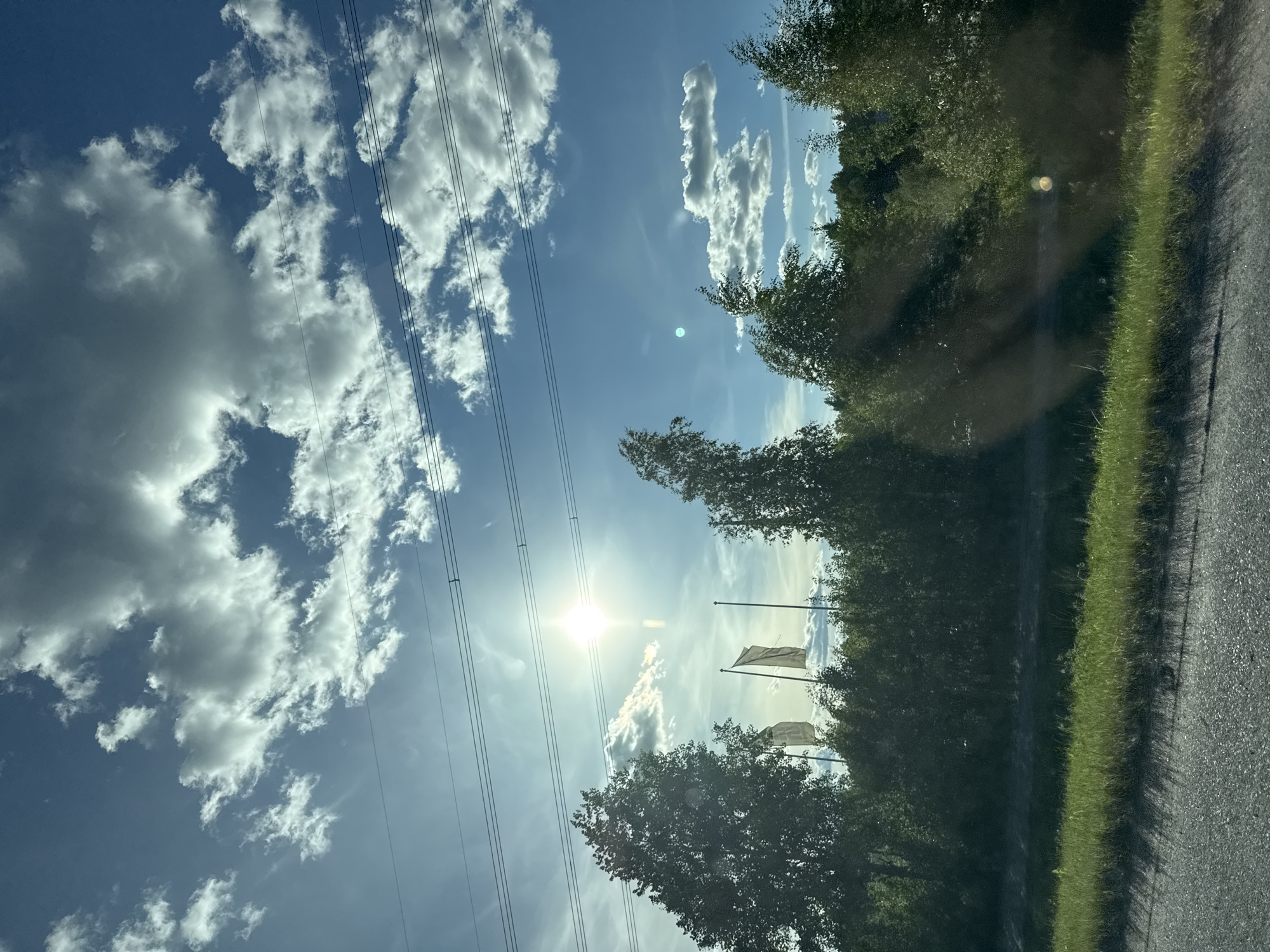 The image captures a bright, sunny day with a vibrant blue sky filled with dramatic white cumulus clouds. Power lines are visible across the upper portion of the frame. In the middle ground, a line of lush green trees forms a dense foliage border. Among the trees, two plain, light-colored flags on poles are visible, their tops barely clearing the tree line. The sun is a brilliant, starburst-like point of light, partially obscured by clouds, casting a strong glare. The foreground shows a strip of green grass along the edge of an asphalt road, with shadows from the trees cast across it. The scene appears to be an outdoor, rural or semi-rural setting, possibly along a road.