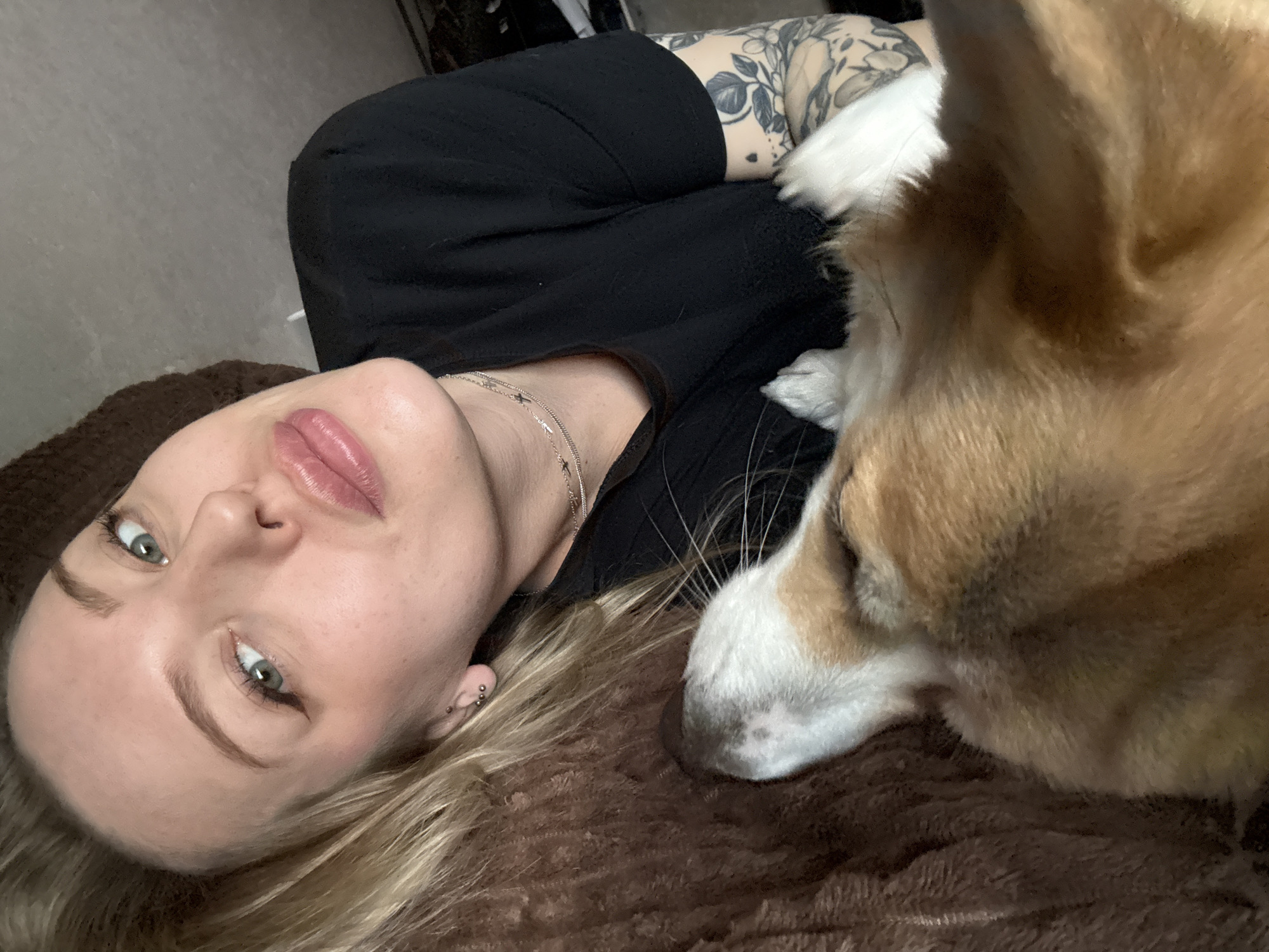 The image captures a close-up, high-angle shot of a young woman and a dog resting together indoors. The woman, with blonde hair, fair skin, and light-colored eyes (likely green or hazel), is lying on her back, looking directly towards the camera. She has full lips with a pinkish tint, and her expression is relaxed and calm. She is wearing a black top and a delicate silver necklace with small charms. Her right arm, visible from the bicep upwards, is adorned with a detailed floral tattoo. On her visible left ear, she has multiple silver stud piercings.
Beside her, on the right side of the frame, is a dog with tan/brown and white fur, resembling a Corgi or a similar breed. The dog's head is resting near the woman, looking away from the camera, appearing peaceful and relaxed. Both the woman and the dog are lying on a soft, brown, textured blanket or bed covering, suggesting a cozy indoor setting. The overall mood of the image is one of companionship, comfort, and tranquility. No specific time of day, weather conditions, or visible text can be discerned from the image, aside from the general indoor location. The location context provided is Borzya, Russia.