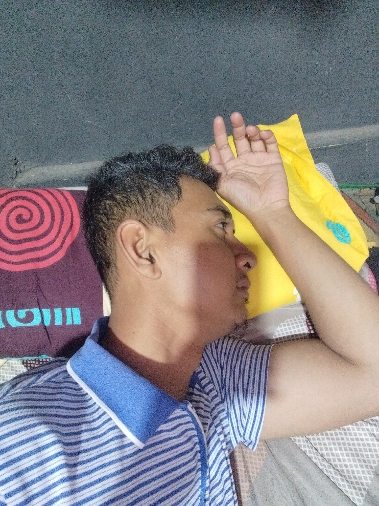 A man is lying down, resting his head on his arm, which is propped up by a yellow pillow. He is wearing a blue and white striped polo shirt. A pillow with a red spiral pattern is visible to his left. The background appears to be a textured wall. The scene is indoors, suggesting a bedroom or living space. This image was taken in Cemplang, Indonesia.