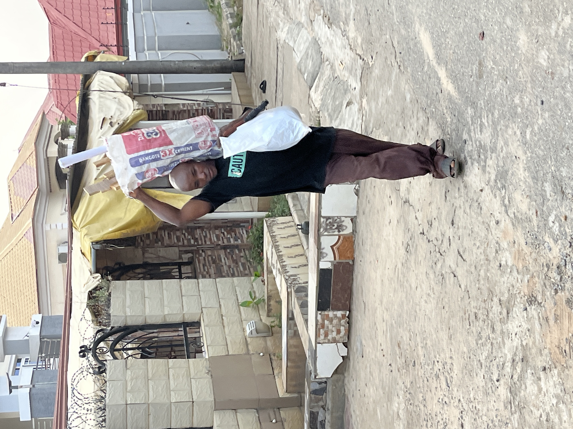 A man is carrying construction materials in an urban environment in Abuja, Nigeria. The man, wearing a black shirt with green text and brown pants, is carrying a bag of cement on his head with wooden planks. He also has a white plastic bag on his shoulder. The background shows buildings with barbed wire fencing and decorative metalwork, suggesting a residential or commercial area. The ground is a concrete surface with visible cracks and wear. Overhead power lines are also visible.