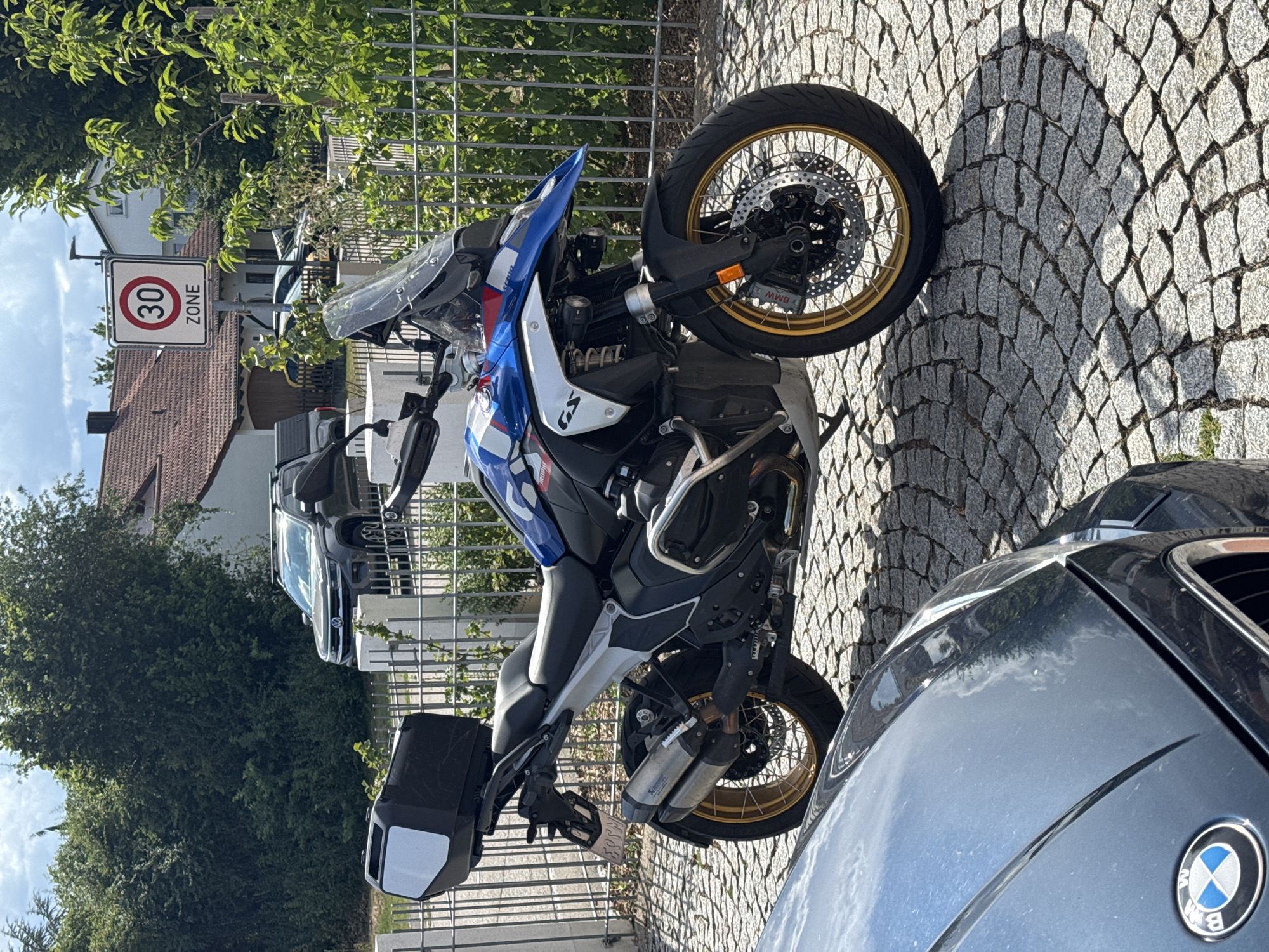 The image depicts a BMW R1250GS Trophy motorcycle parked on a cobblestone street in Wettstetten, Germany, near a fence and some vegetation. A portion of a dark-colored BMW car is visible in the foreground. A speed limit sign indicating "30 ZONE" is posted on a pole behind the motorcycle. In the background, there are houses with tiled roofs and trees. The sky is partly cloudy.