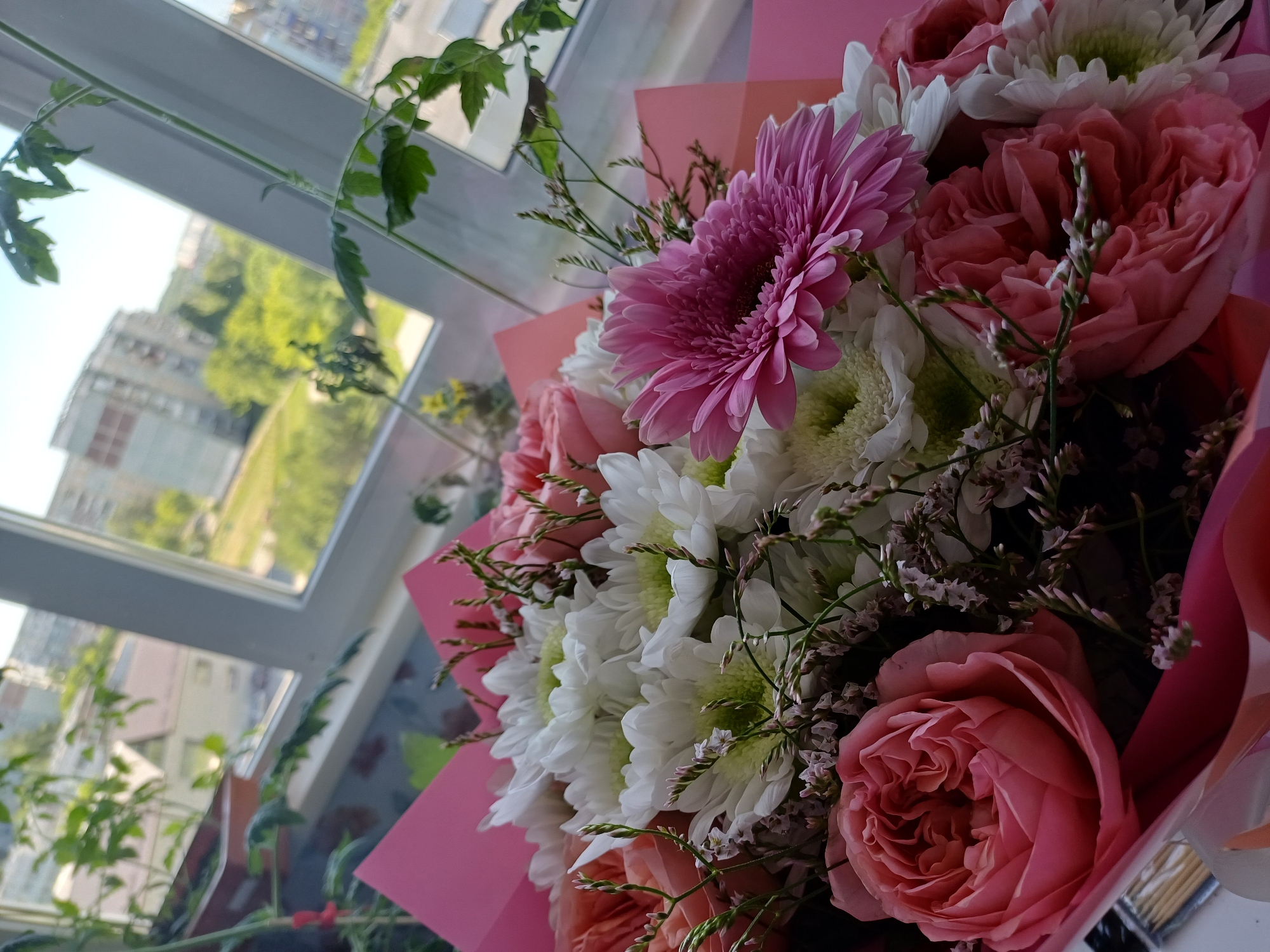 A bouquet of flowers is placed on a windowsill, with a view of a city in the background. The bouquet includes pink roses, white chrysanthemums, and a pink gerbera daisy. The flowers are arranged with greenery and delicate sprigs. Behind the bouquet, a window shows buildings, trees, and a bright sky, suggesting an urban setting. The overall scene appears to be indoors, by a window, likely in a residential or office space.