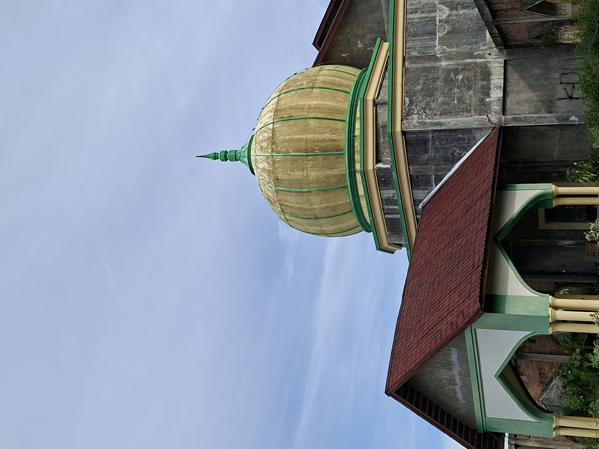A low-angle, slightly tilted view of a building in Lhokseumawe, Indonesia, highlights architectural elements. The building exhibits a yellow dome with green outlining, topped with a pointed green spire. Attached to the dome is a gray concrete structure. Adjacent to the gray structure, there is a roof with a red tiled surface. A section of the building with green and white detailing is visible, featuring yellowish pillars. The background reveals a blue sky with light clouds. The overall impression is of an architectural portrait, focusing on the building's unique details.
