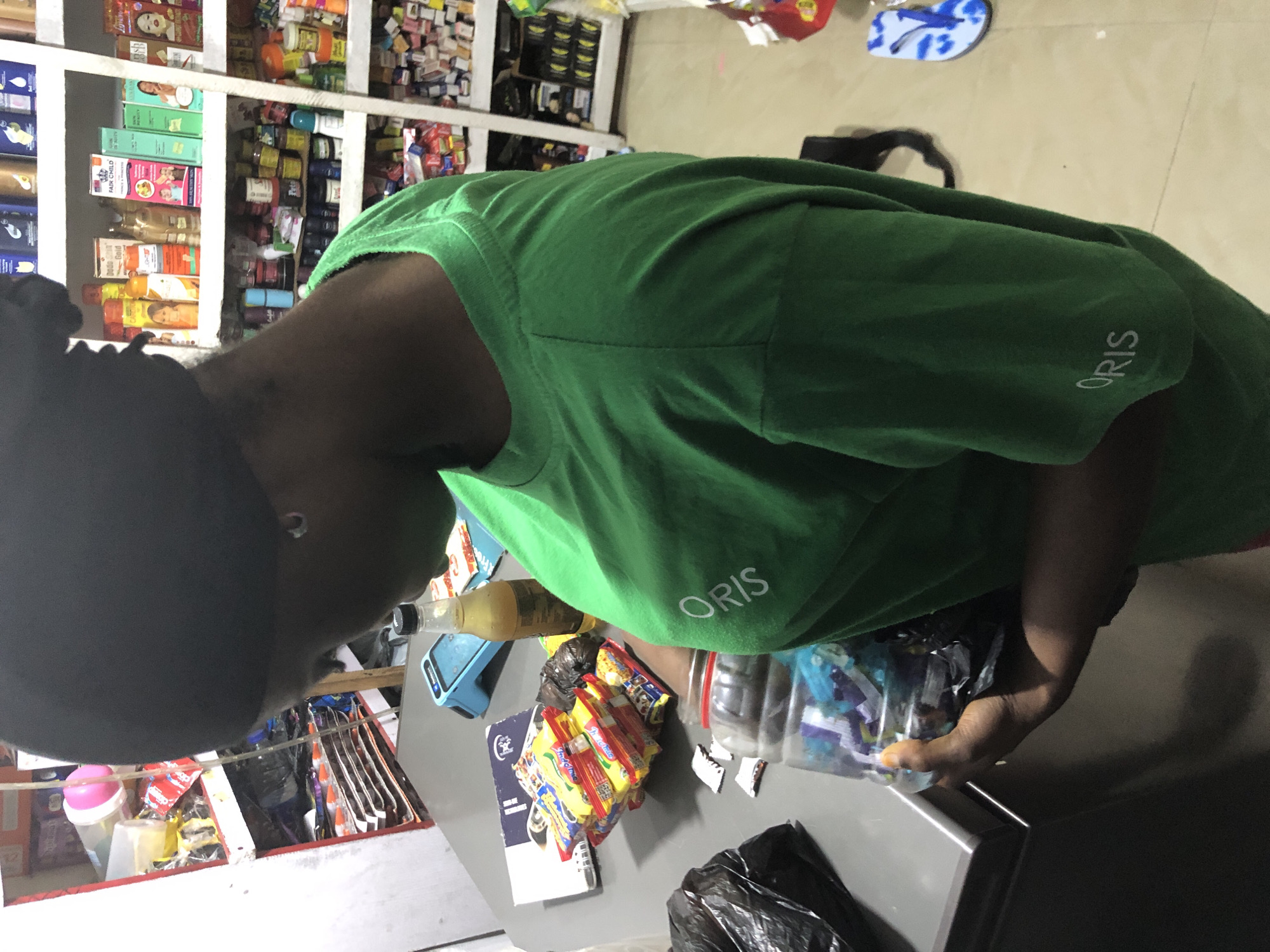 The image shows a young Black person wearing a green t-shirt with the word "ORIS" printed on it in white. They are standing in what appears to be a small shop or market stall in Ado, Nigeria. The person is holding a clear plastic bottle filled with colorful items, possibly candy wrappers or small toys. In front of them is a counter with a stack of packaged snacks, a blue card reader, and a bottle of yellow liquid. Behind them, shelves are stocked with various products, including toiletries, food items, and cosmetics. The overall scene suggests a retail environment.