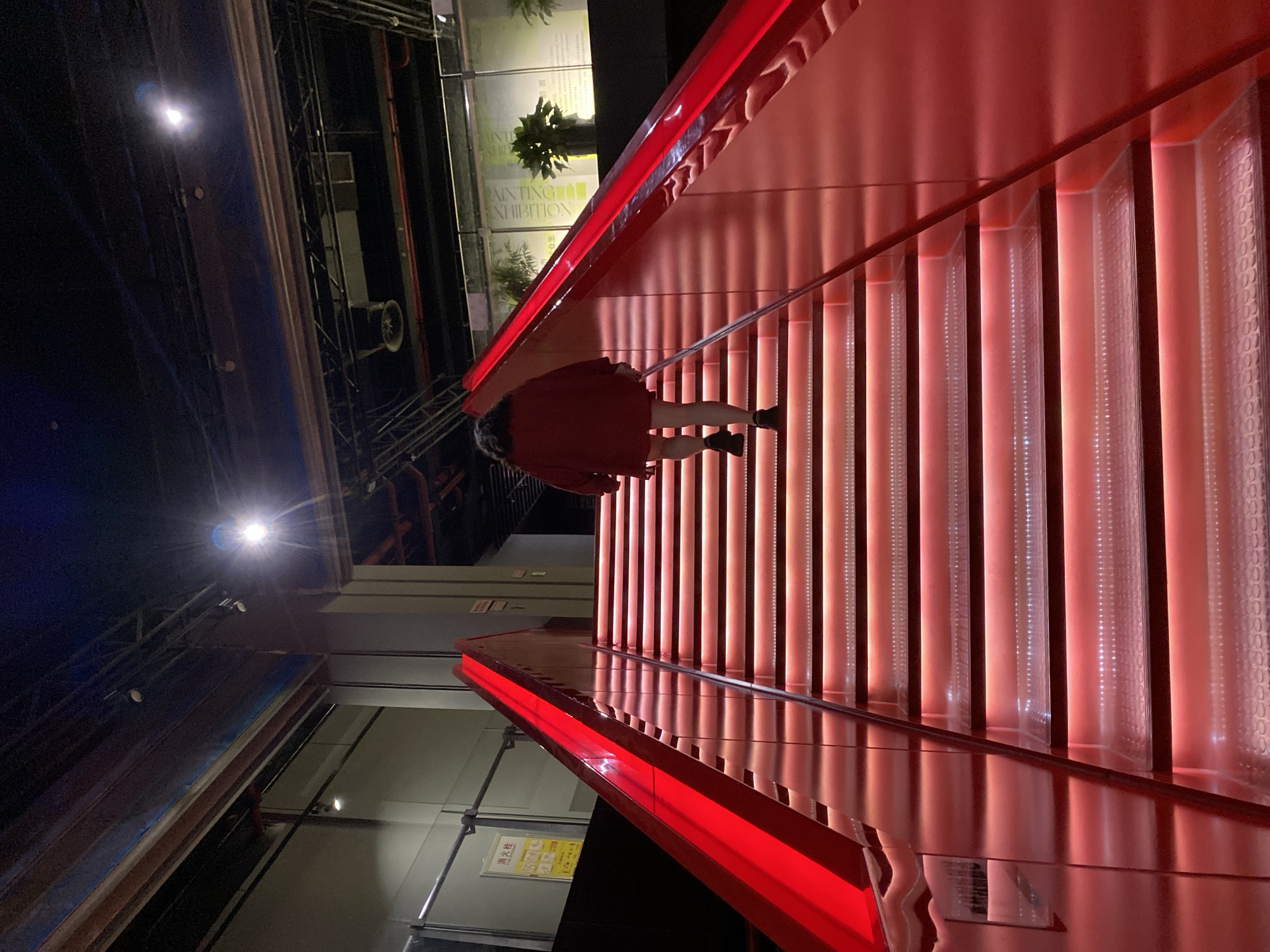 In Yuzhong District, China, a person, viewed from behind, walks up a brightly lit, illuminated staircase. The staircase, with red illuminated steps, ascends diagonally from the bottom left to the top right of the frame. The person is wearing a red jacket and dark shorts, with dark shoes. The surrounding environment appears to be an indoor public space, possibly a museum or exhibition hall, with exposed industrial-style ceiling elements, lights, and signage visible in the background. The lighting is dramatic, with the red glow from the stairs dominating the scene.