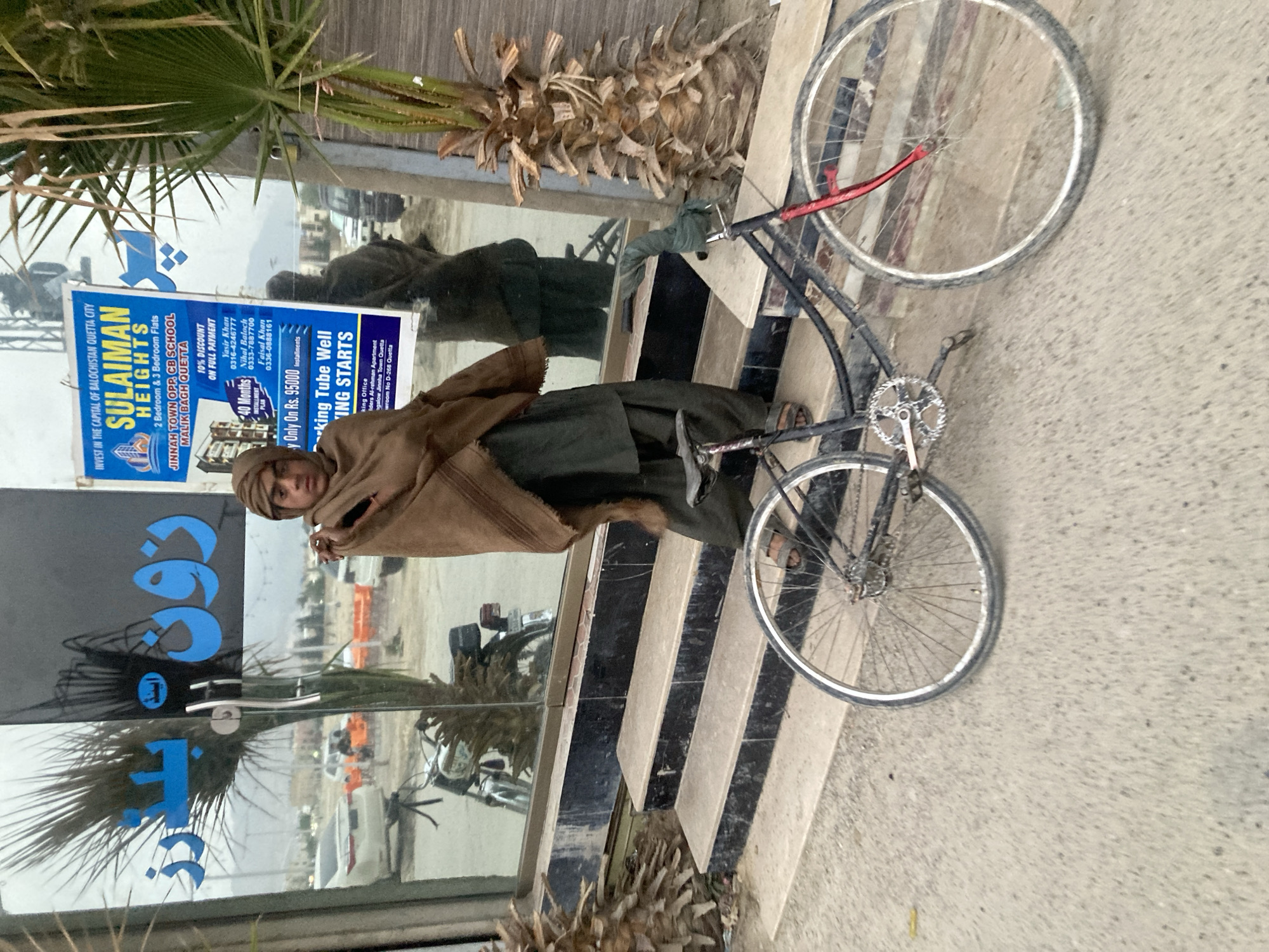 A man stands outside a building in Quetta City Tehsil, Pakistan, next to a bicycle. A promotional sign for "Sulaiman Heights" is visible, advertising apartments in Jinnah Town, opposite CB School in Malik Bagh, Quetta. The building has mirrored windows. A palm tree is near the building.