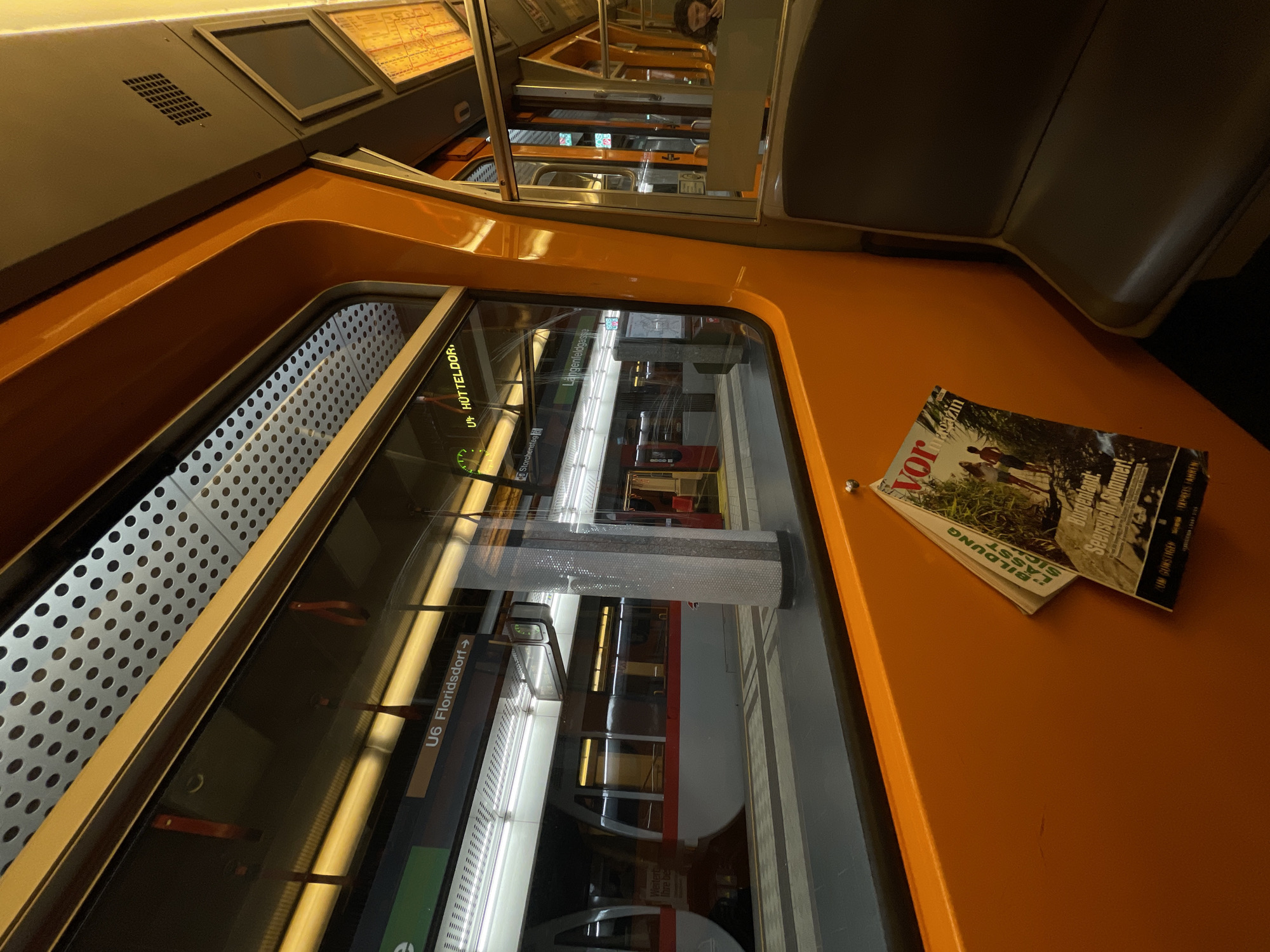The image shows the interior of a U6 subway train in Vienna, Austria. It's taken from a high angle, focusing on the orange interior of the train. A "VOR" magazine lies on the seat. The subway line U6 is indicated with "Floridsdorf" being the next station. The reflection in the window shows another train at a station, suggesting the photo was taken during a stop. "Hütteldorf" and "Langenfeldgasse" are also visible on signs within the train, marking other stops on the U6 line.