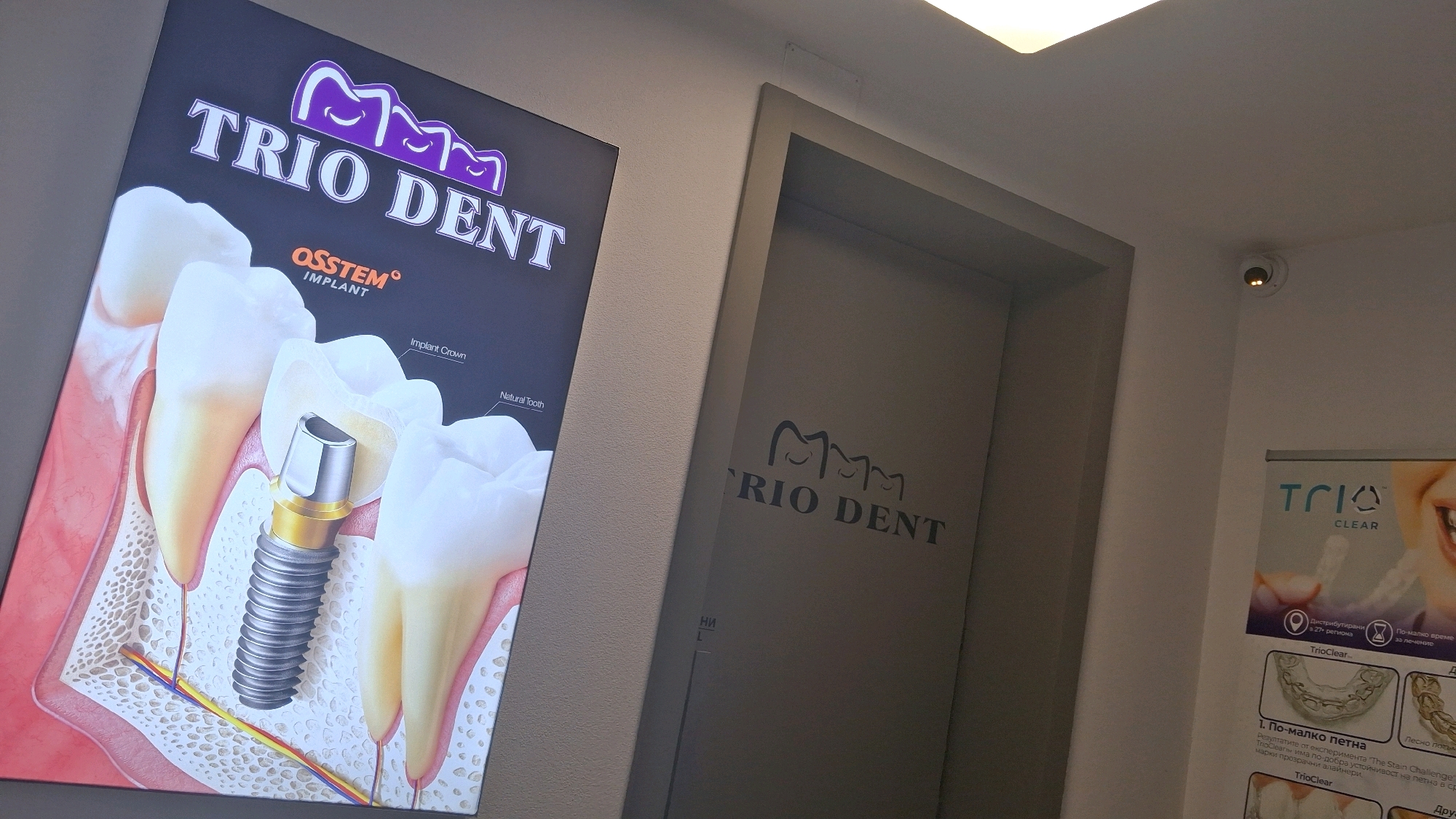 The image shows the interior of a dental clinic in Gotse Delchev, Bulgaria. The focal point is a large illuminated advertisement for "TRIO DENT" and "OSSTEM IMPLANT", featuring a detailed anatomical illustration of a dental implant and tooth. To the right of the advertisement, there is a door or entrance to another area, also branded with "TRIO DENT" in a darker, debossed style. Further to the right, a banner displays information about "TRIO Clear" aligners and a smiling person's teeth. A security camera is visible on the upper right portion of the wall, near the ceiling. The lighting suggests an indoor commercial setting.