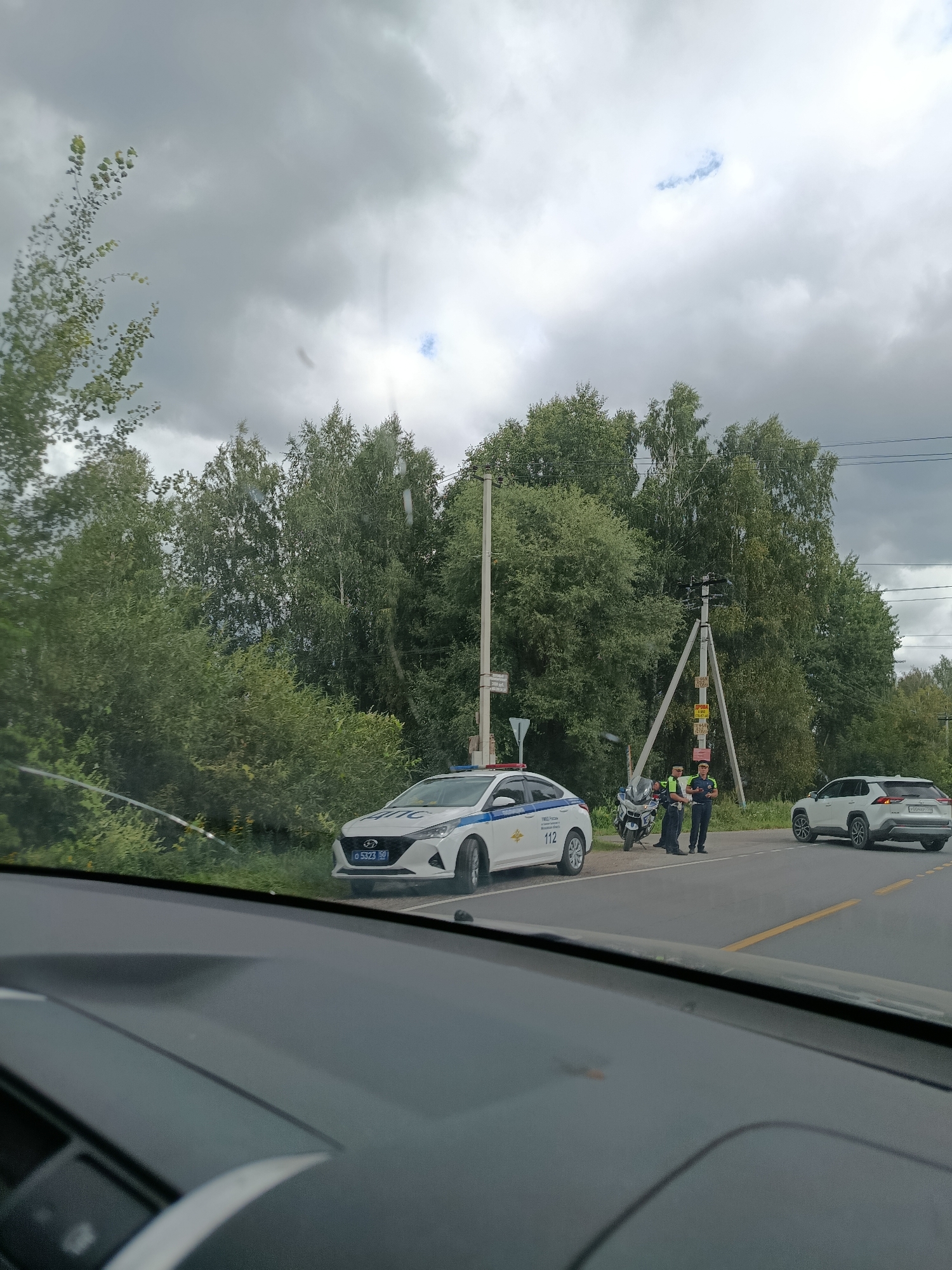 The image depicts a roadside scene, possibly in or near 1-Go Maya, Russia. The sky is overcast with a mix of dark and lighter clouds. On the asphalt road, a white police car with blue and red markings is parked, displaying the number "112" on its rear. Next to the police car is a motorcycle. Two individuals, appearing to be police officers in uniform, are standing near the motorcycle and a utility pole with wires. A white SUV is visible further down the road on the right. The surrounding environment is characterized by lush green trees and foliage. The perspective suggests the photo was taken from inside another vehicle.